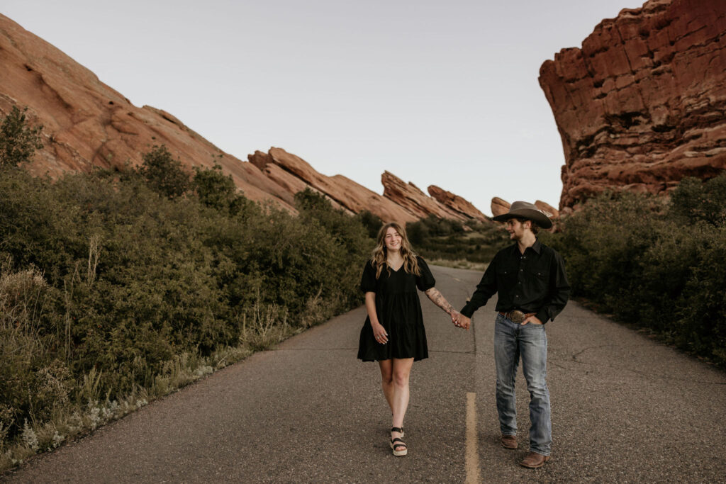 man and woman walk along road during couples portraits at red rocks park.