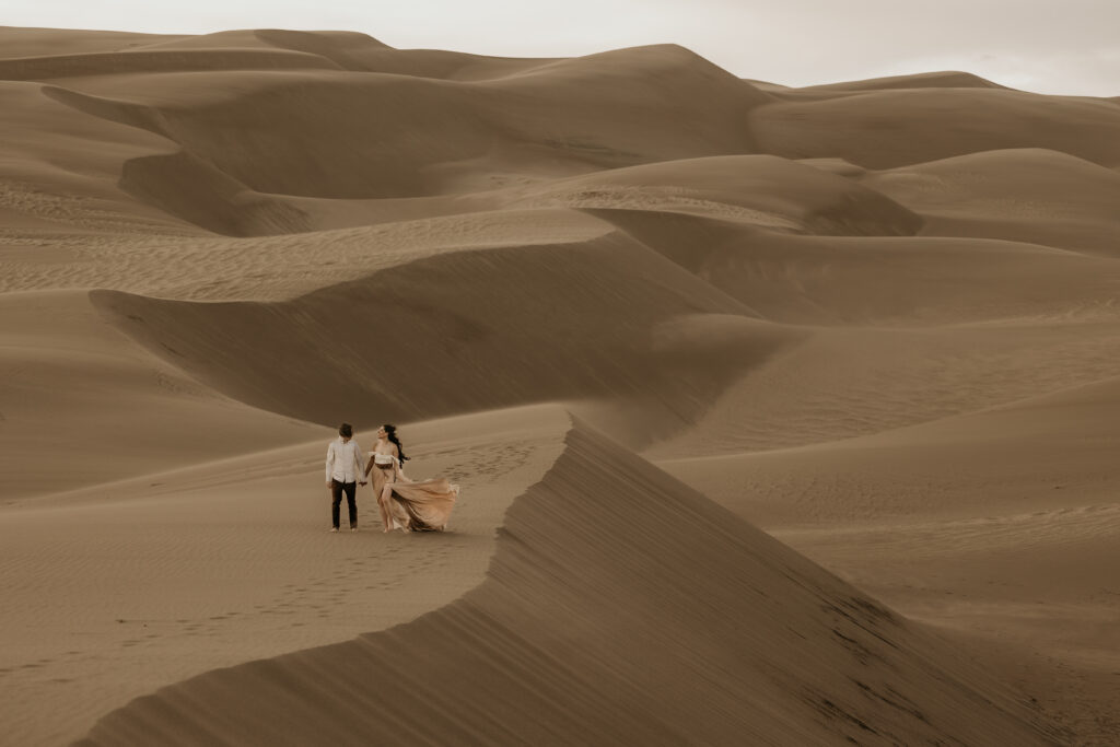 man and woman walk across sand dunes during couples portraits in colorado.