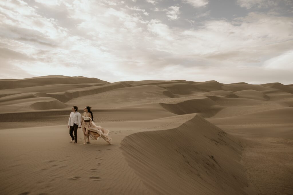 man and woman walk along sand dune during colorado couples portraits.