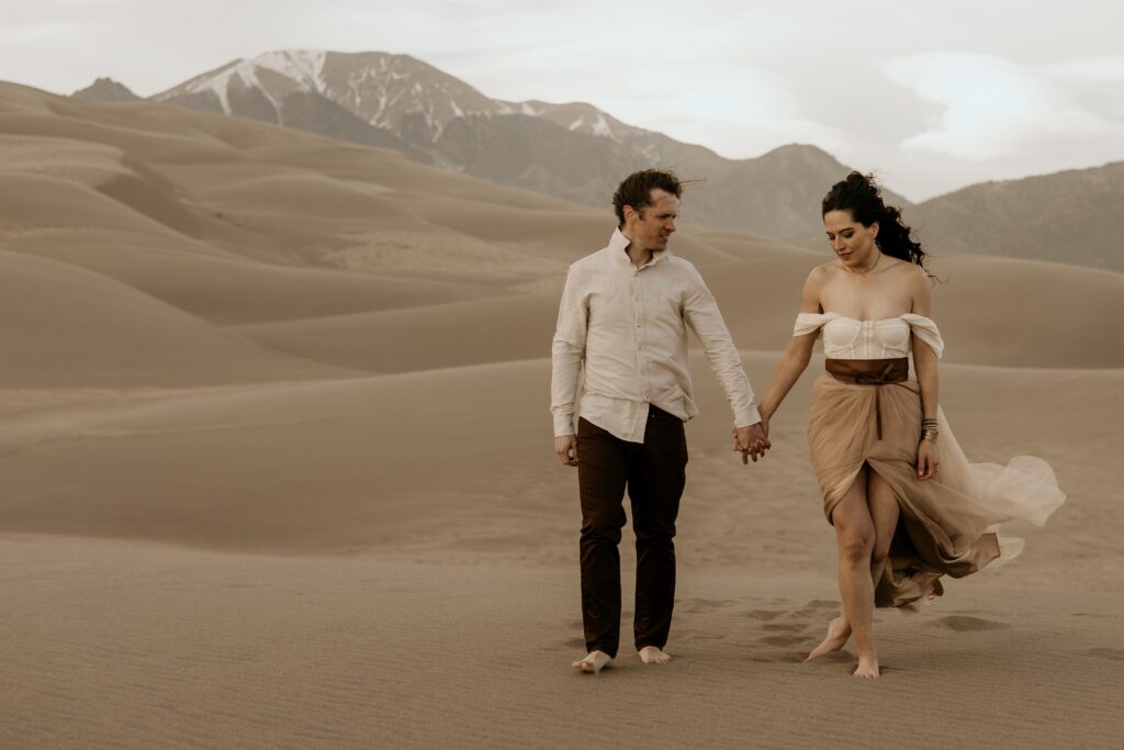 man and woman walk along sand with colorado mountains in the background.