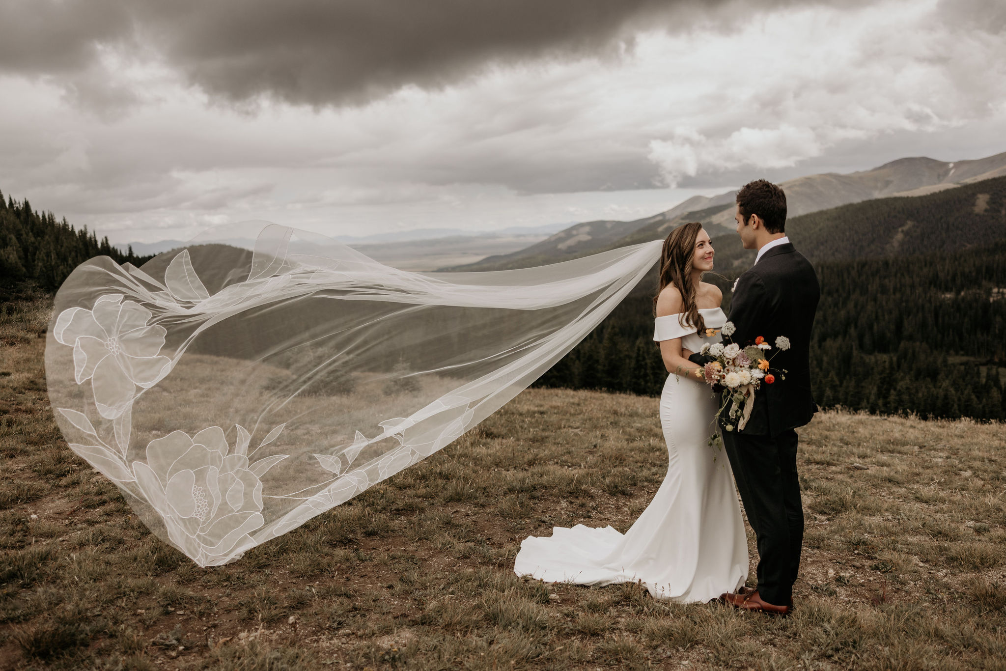 bride and groom stand in front of mountains and look at each other, with wedding veil flowing in the wind.