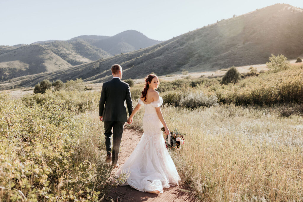 bride and groom walk through field at red rocks park & amphitheatre during wedding portraits.