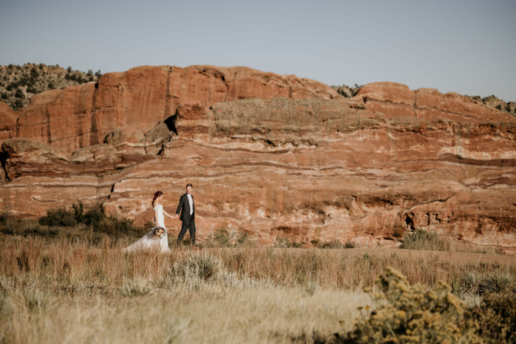 bride and groom walk in front of rock formation at red rocks park & amphitheatre.