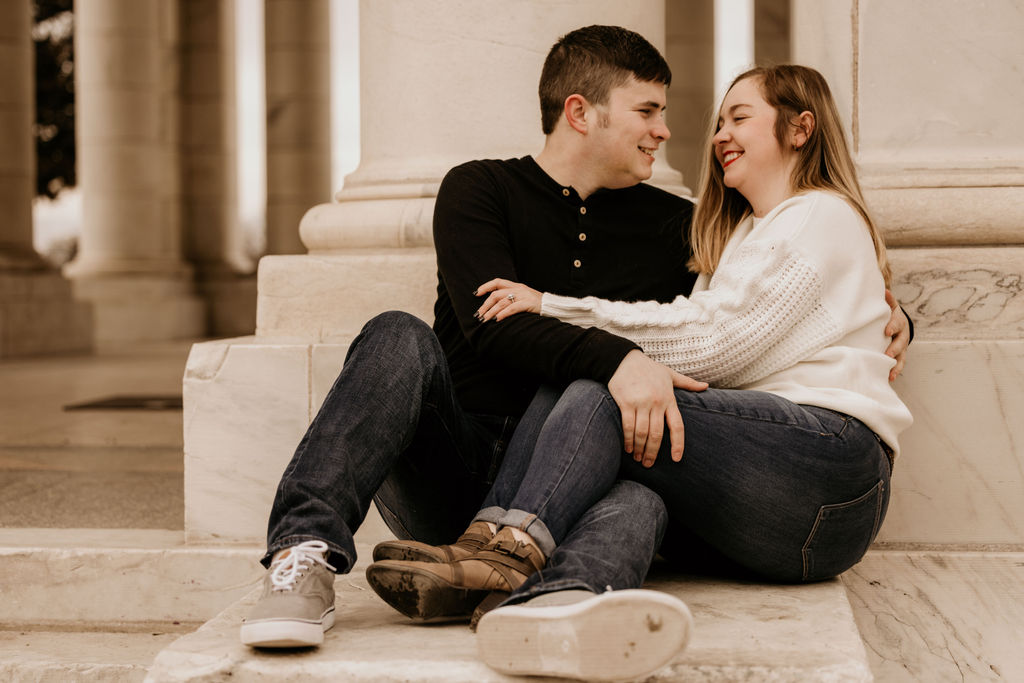 man and woman sit on marble step and smile during engagement photos in denver.
