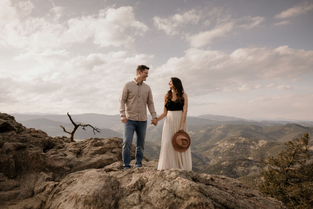 man and woman stand in front of the colorado mountains and smile at each other during engagement photos.