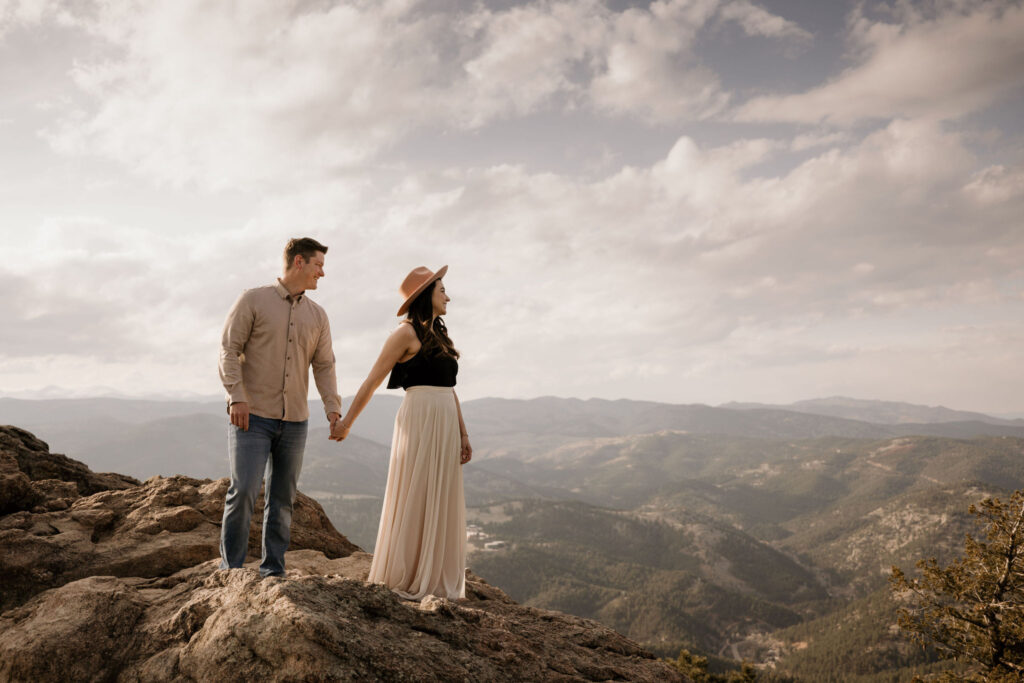 man and woman stand on rock and look at the colorado mountains during engagement photos.