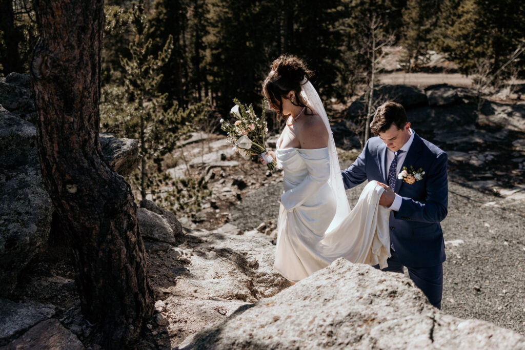 groom holds brides dress while climbing up a rock formation.