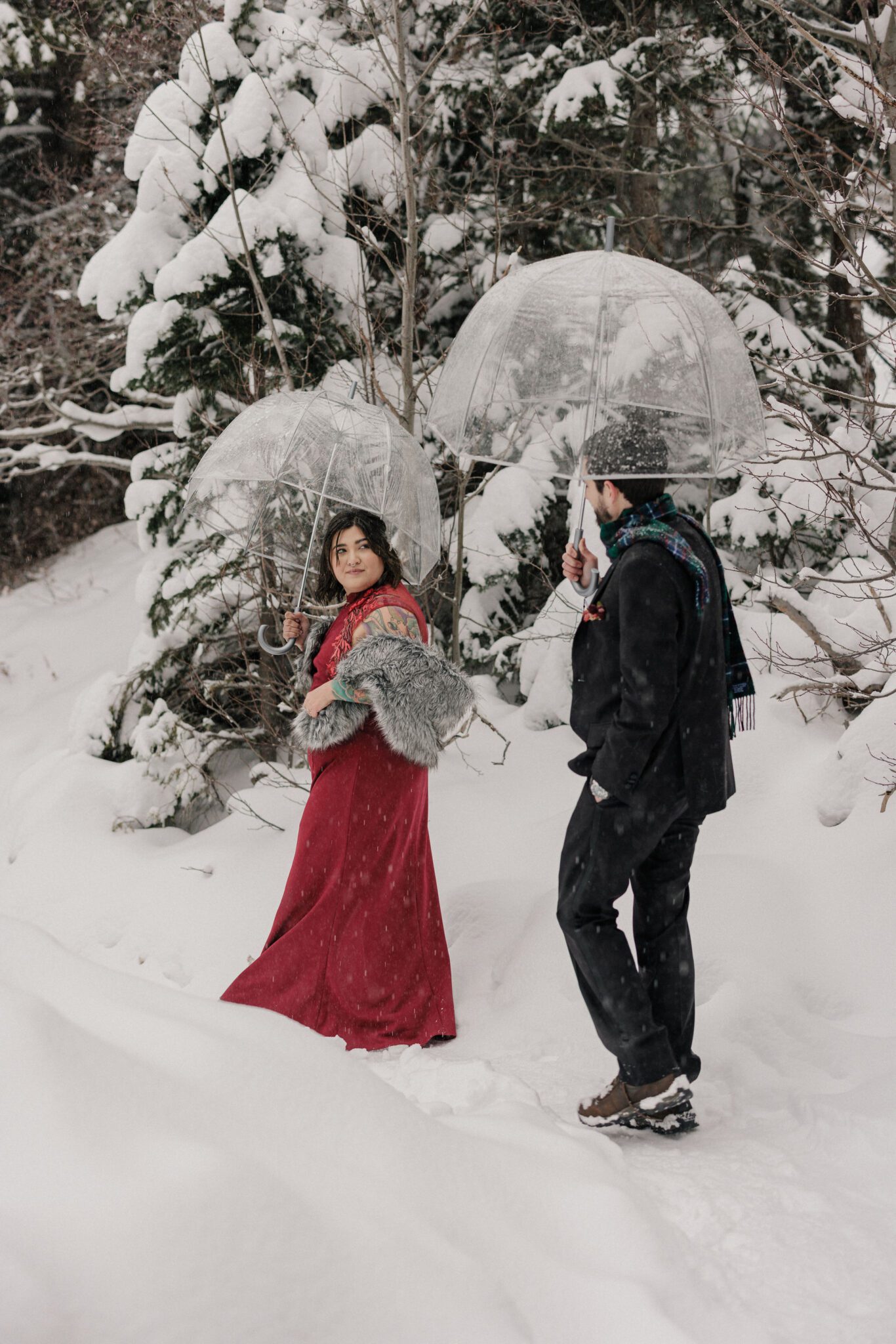 man and woman walk through snow with clear umbrellas during colorado engagement photos