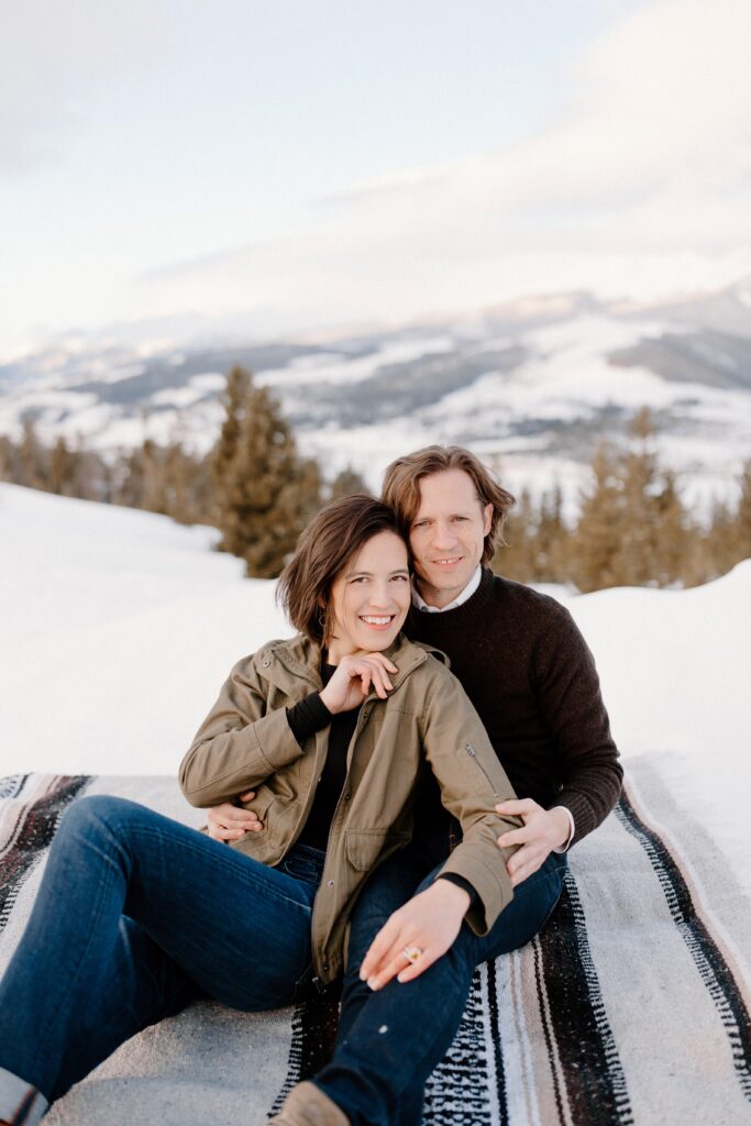 man and woman sit on blanket in the snow during colorado mountain engagement photos.