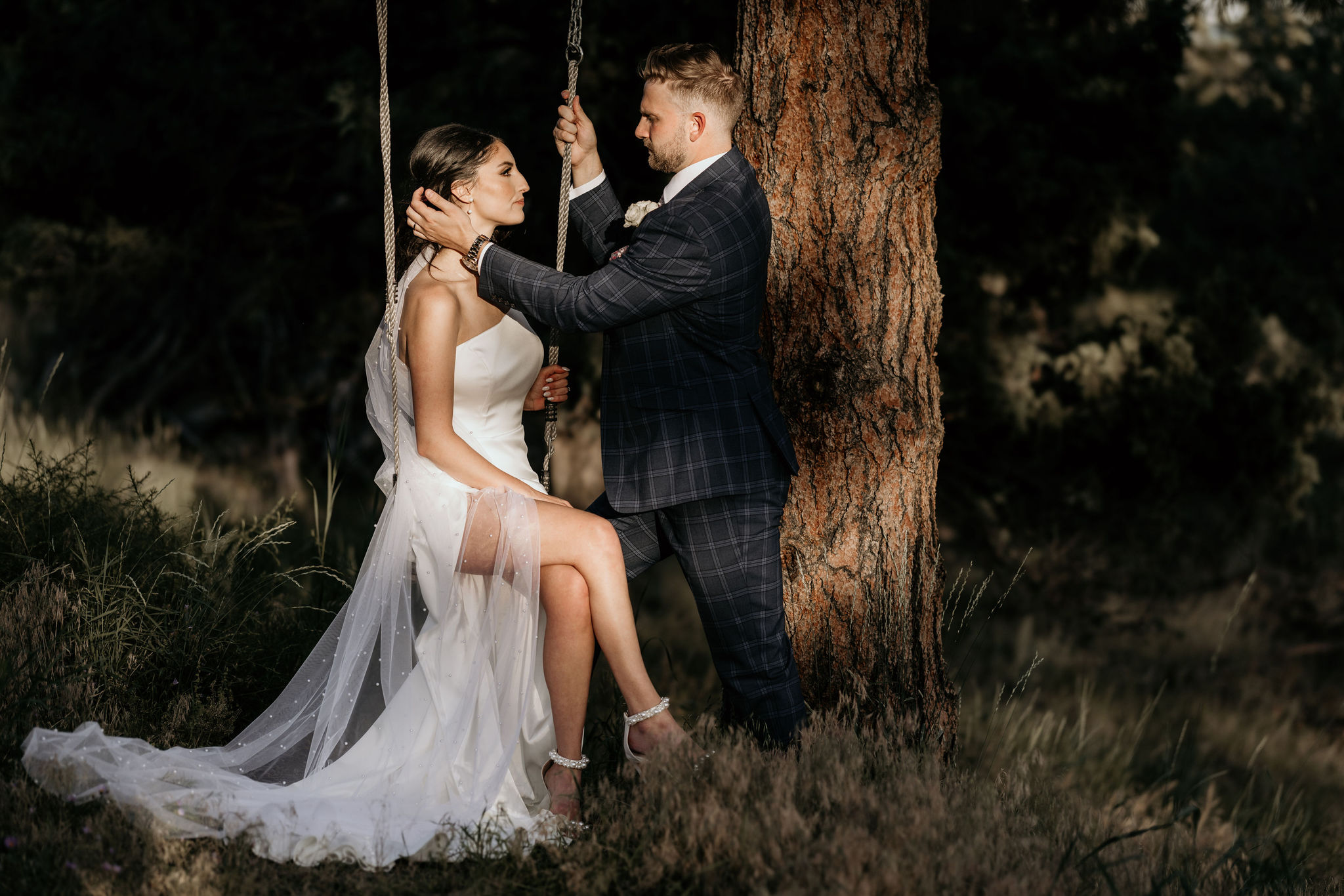 groom puts brides hair behindher ear while she sits on a tree swing.