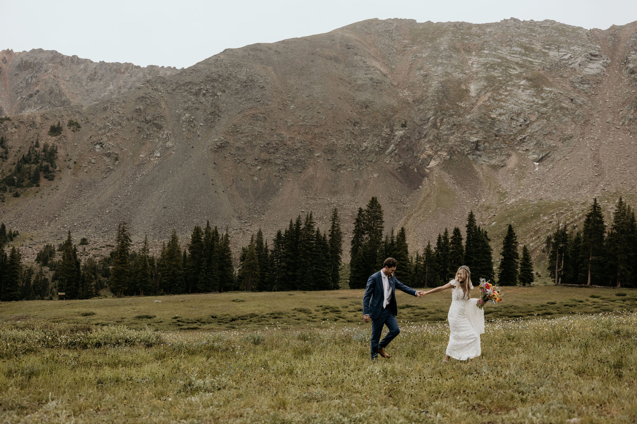 bride and groom hold hands and walk in field in front of colorado mountains