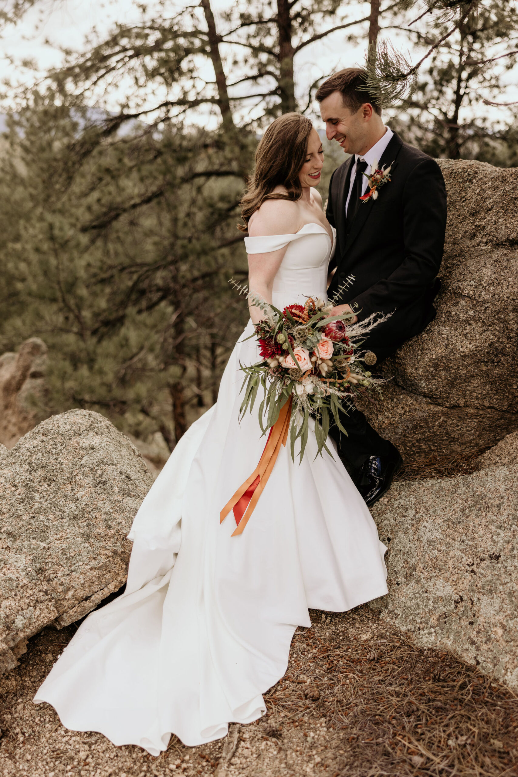 bride and groom lean against a rock and smile during wedding photos.