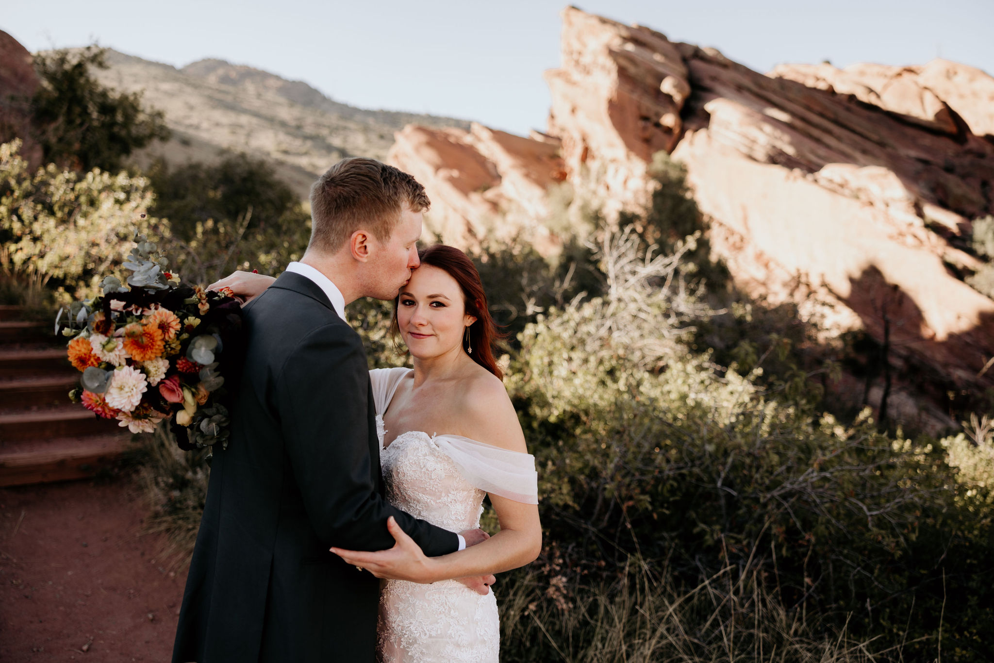groom kisses brides head with towering red rocks in the background.