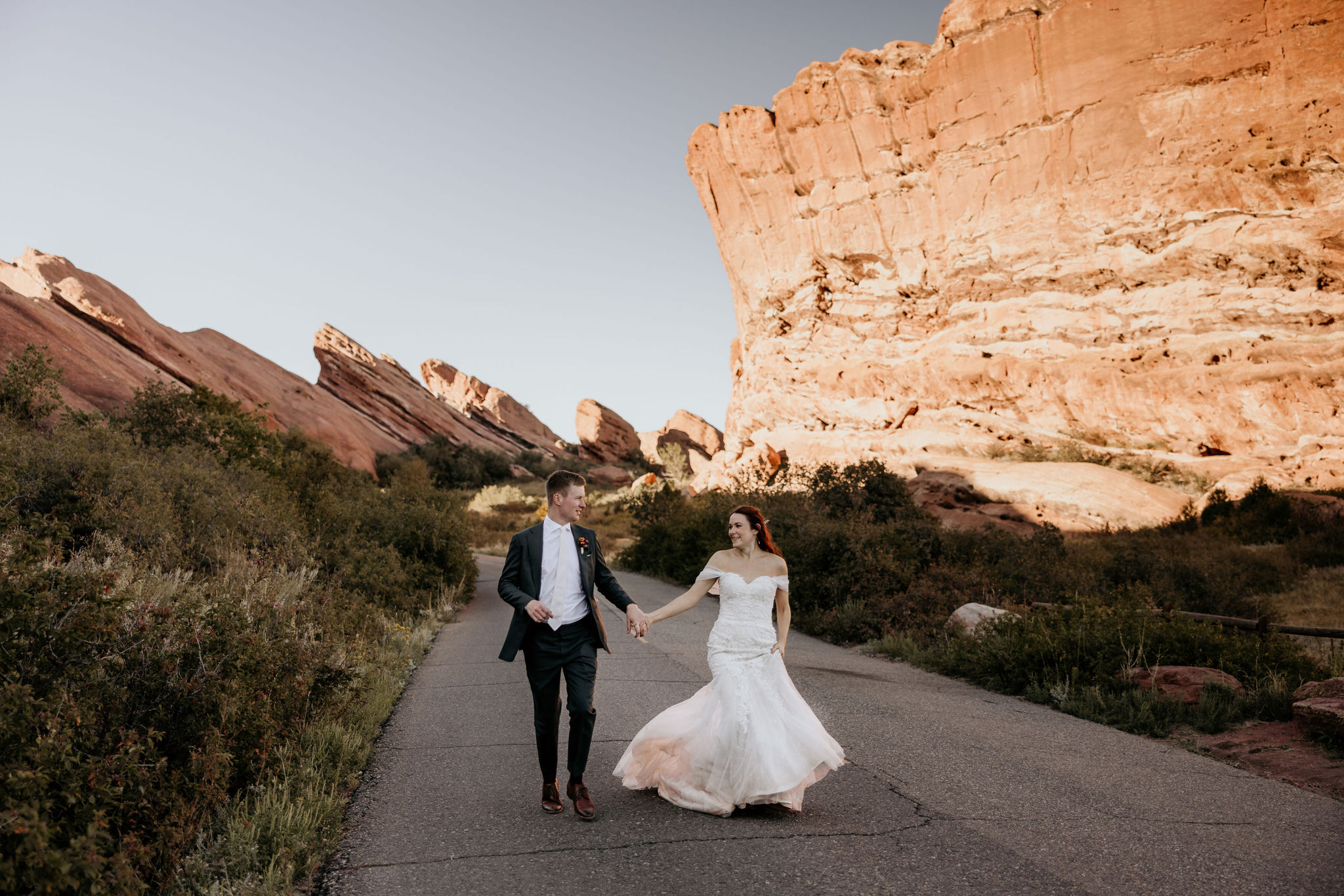 bride and groom hold hands and run down road at red rocks park and amphitheatre in colorado