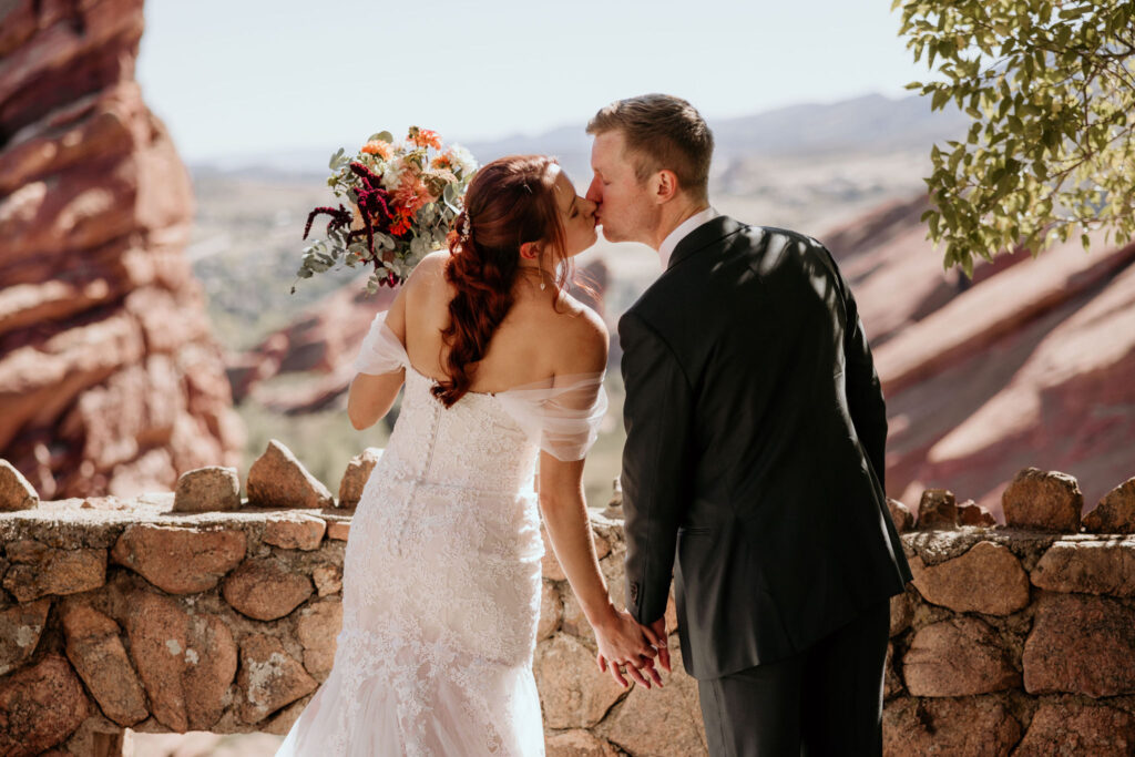 bride and groom hold hands and kiss over the views of red rocks park and amphitheatre.