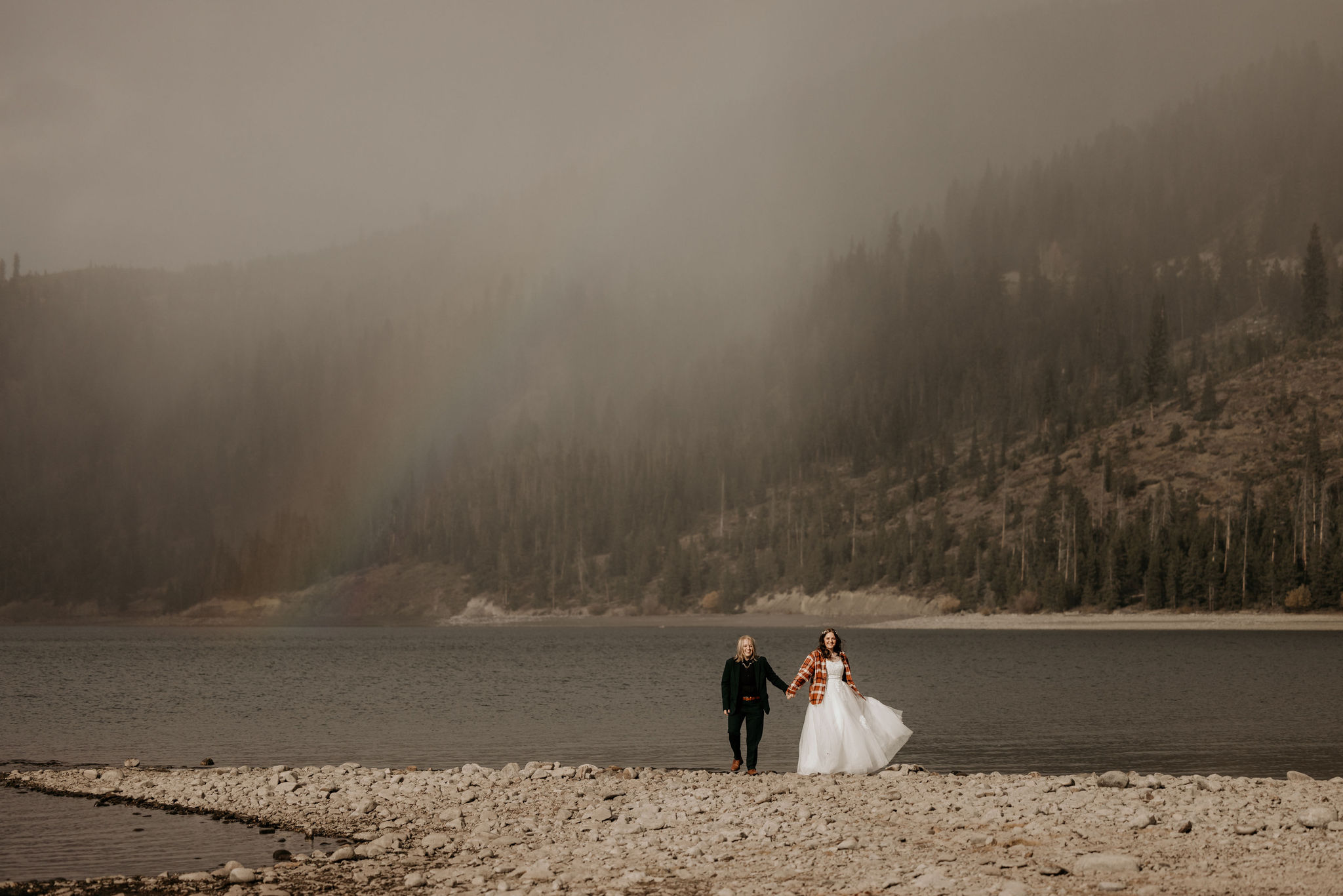 two brides hold hands and walk along colorado lake during elopement.