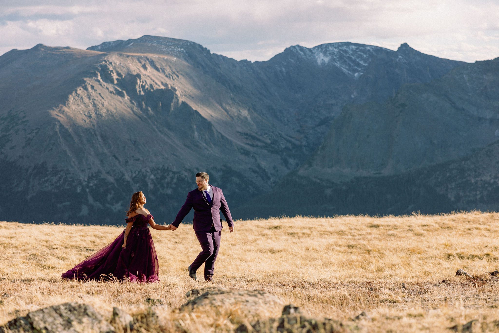 bride and groom, both in purple wedding attire, hold hands and walk in a field in front of the colorado mountains.