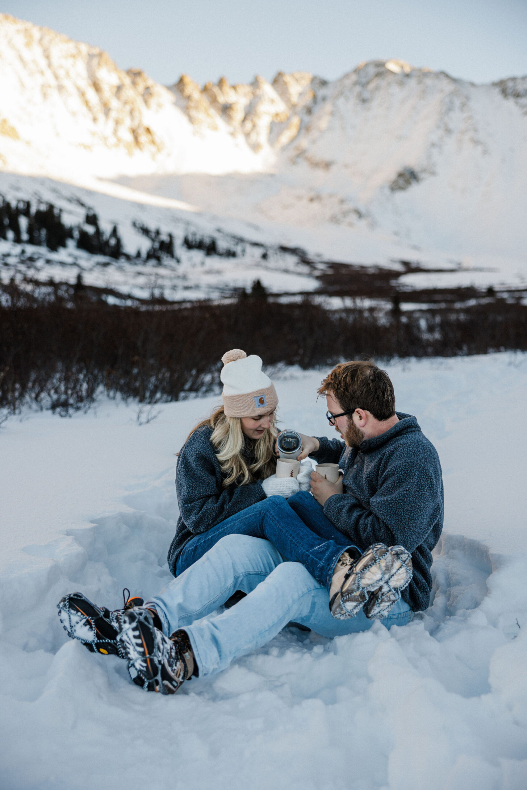 man pours coffee for woman while sitting in the snow during colorado winter engagement photos.