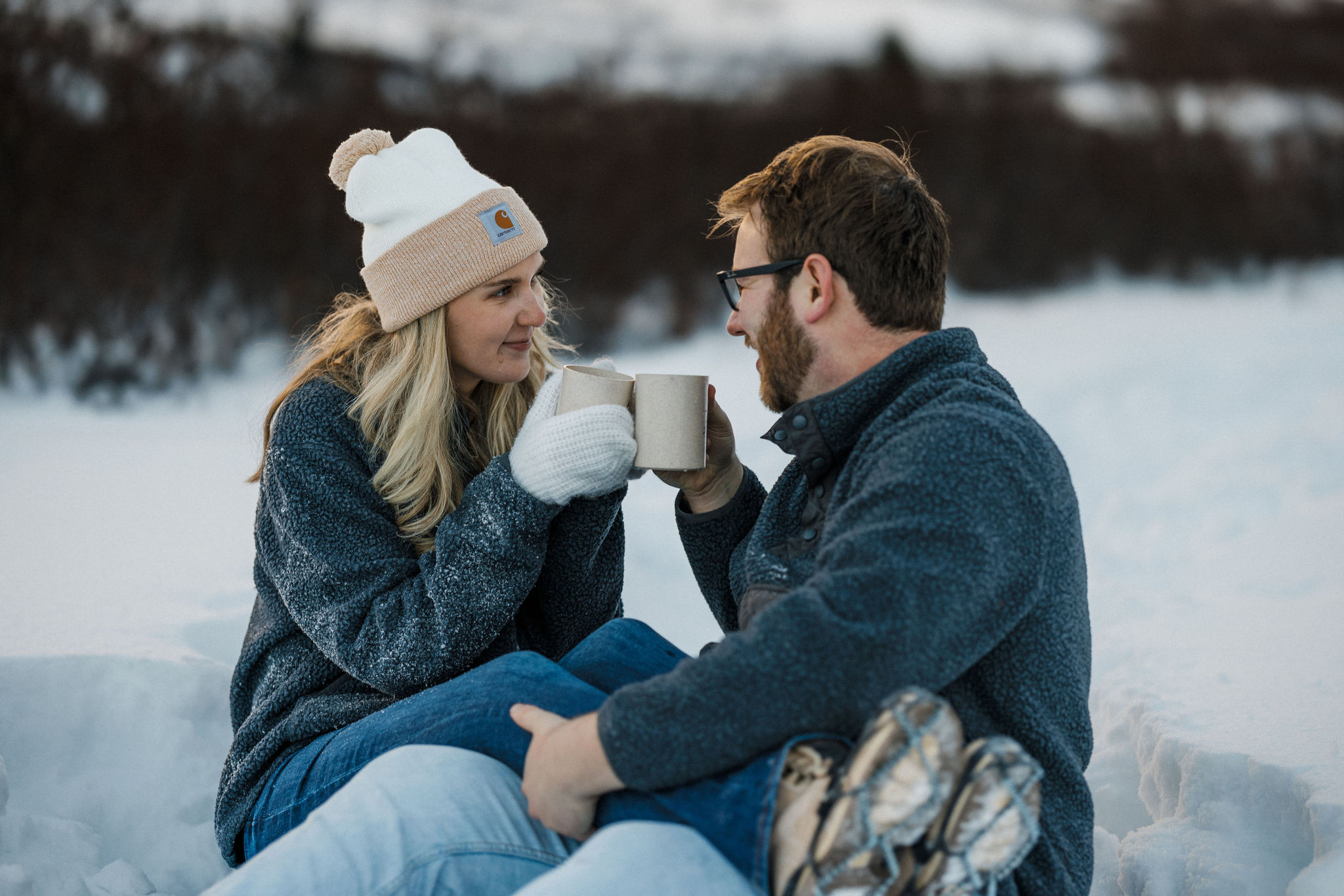 engaged couple cheers mugs while sitting in the snow during winter engagement photos.