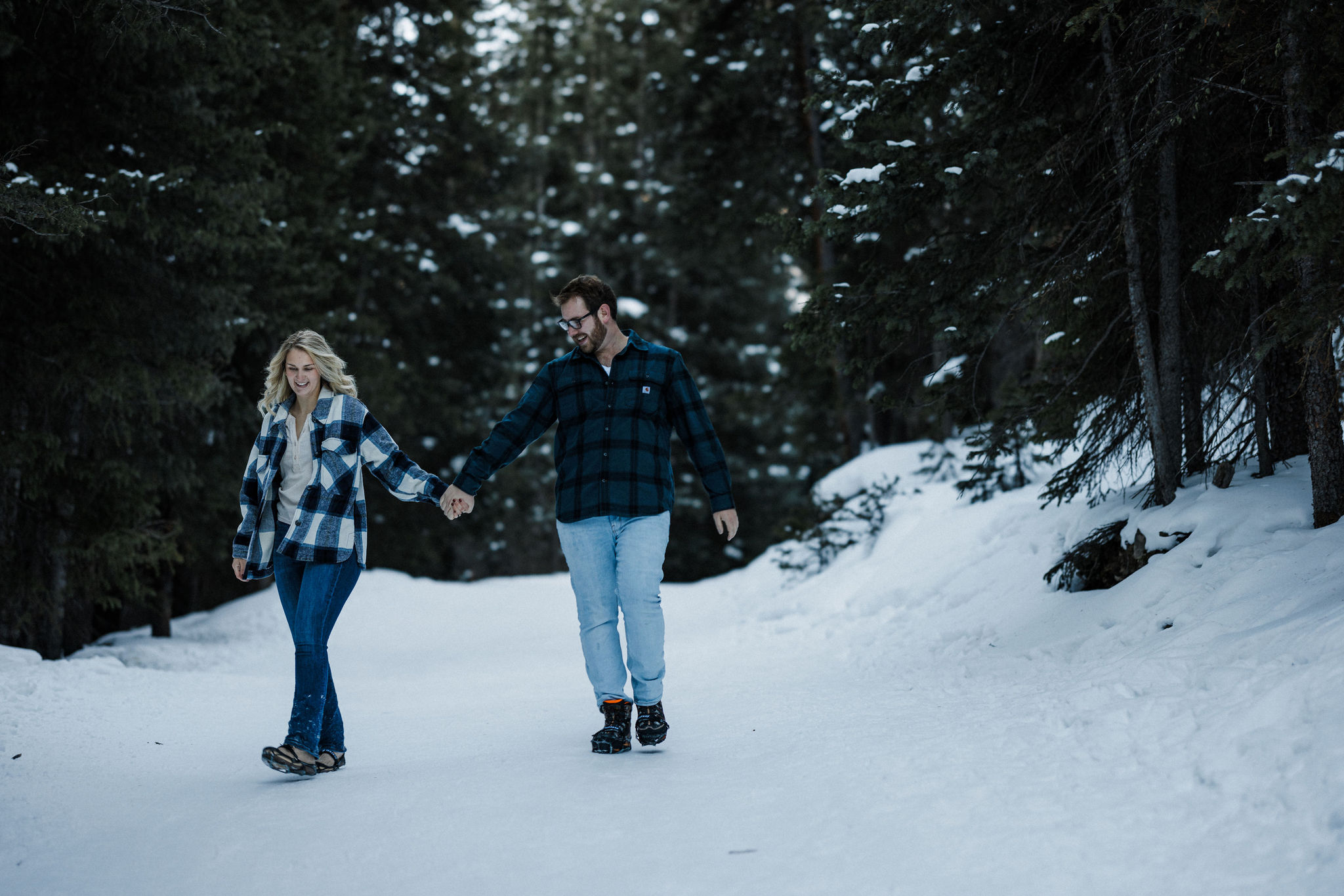 engaged couple hold hands and walk along snowy mountain trail during winter engagement photos