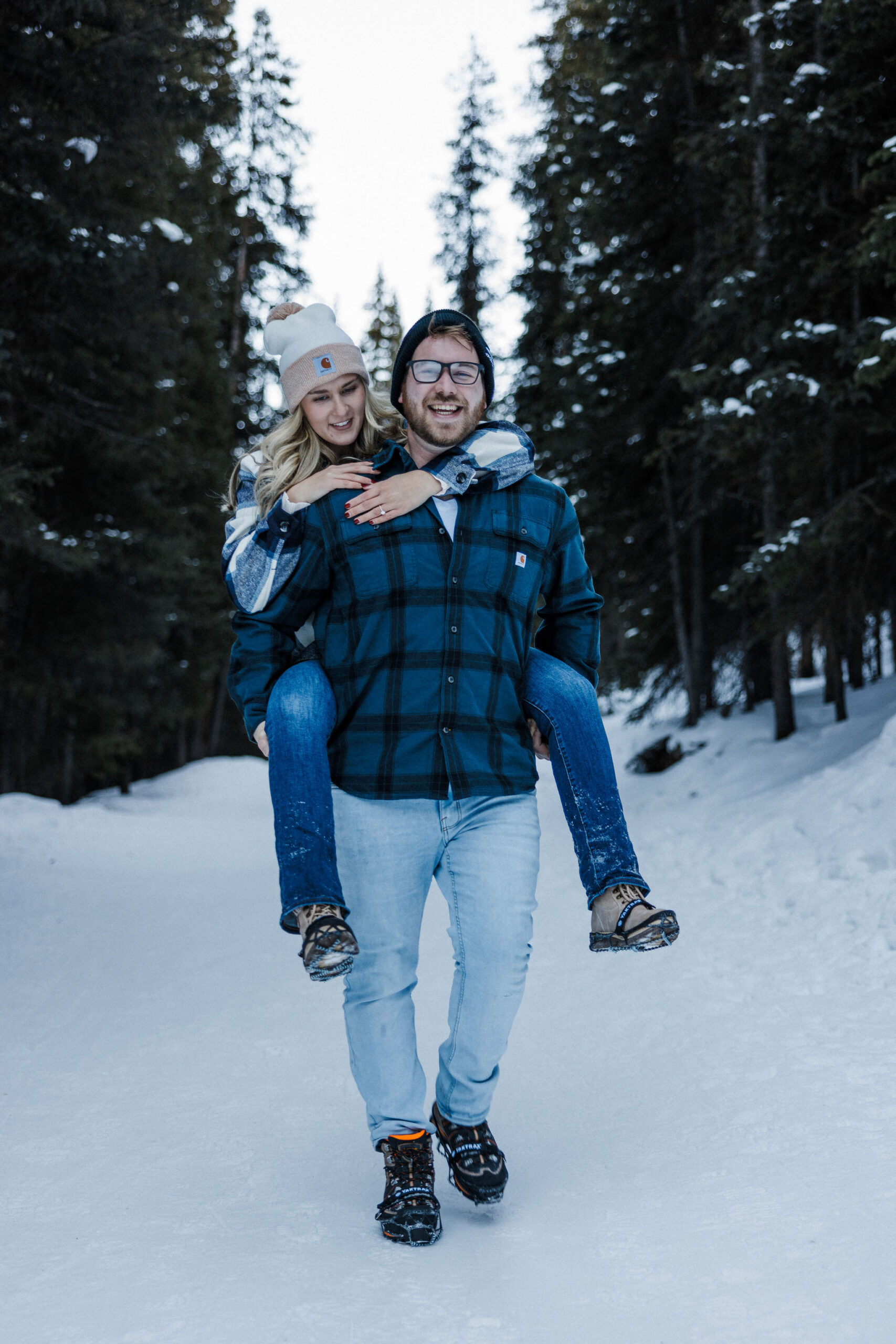 man carries woman on his back during winter engagement photos.