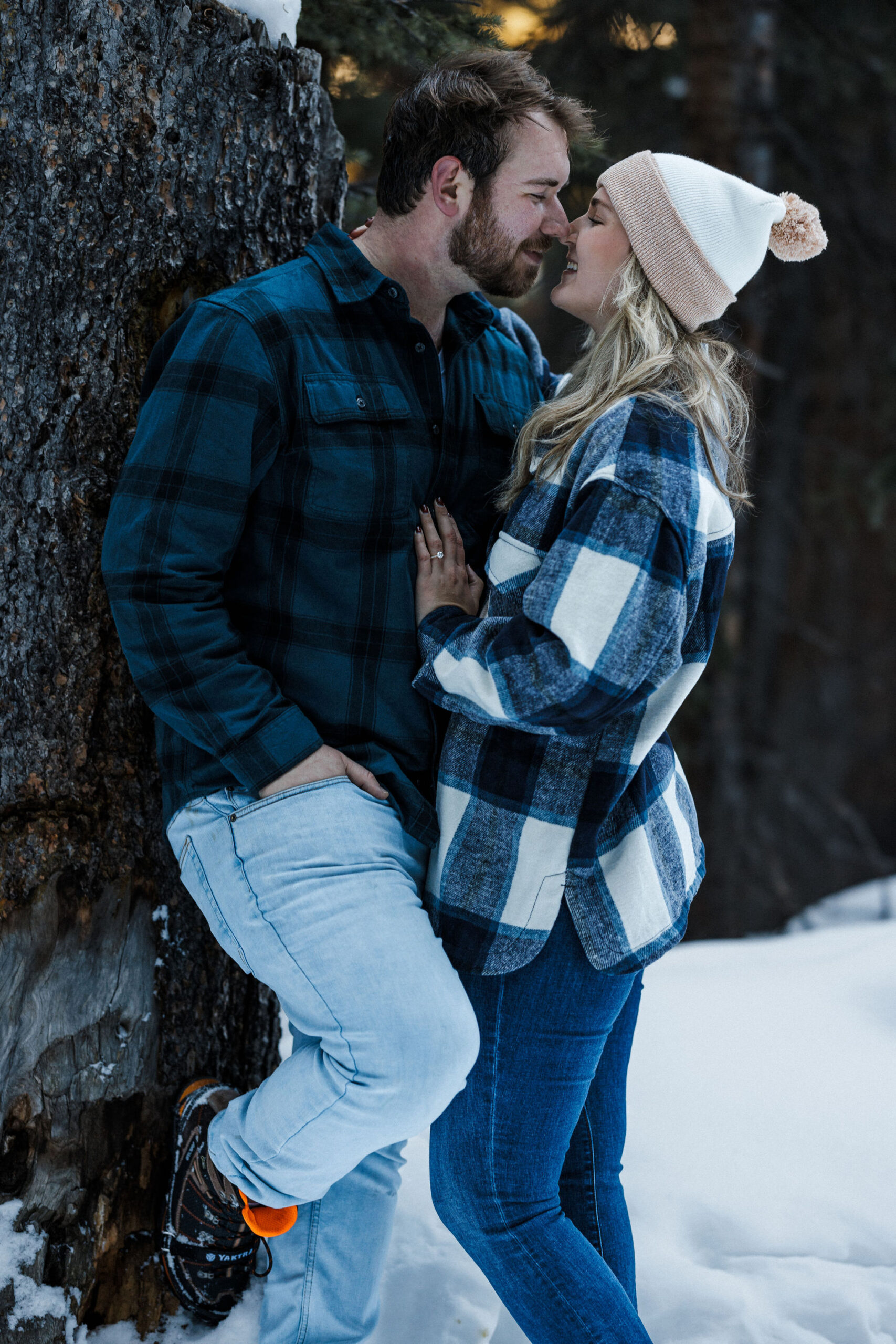 engaged couple leans against tree and goes for a kiss during winter engagement photos.