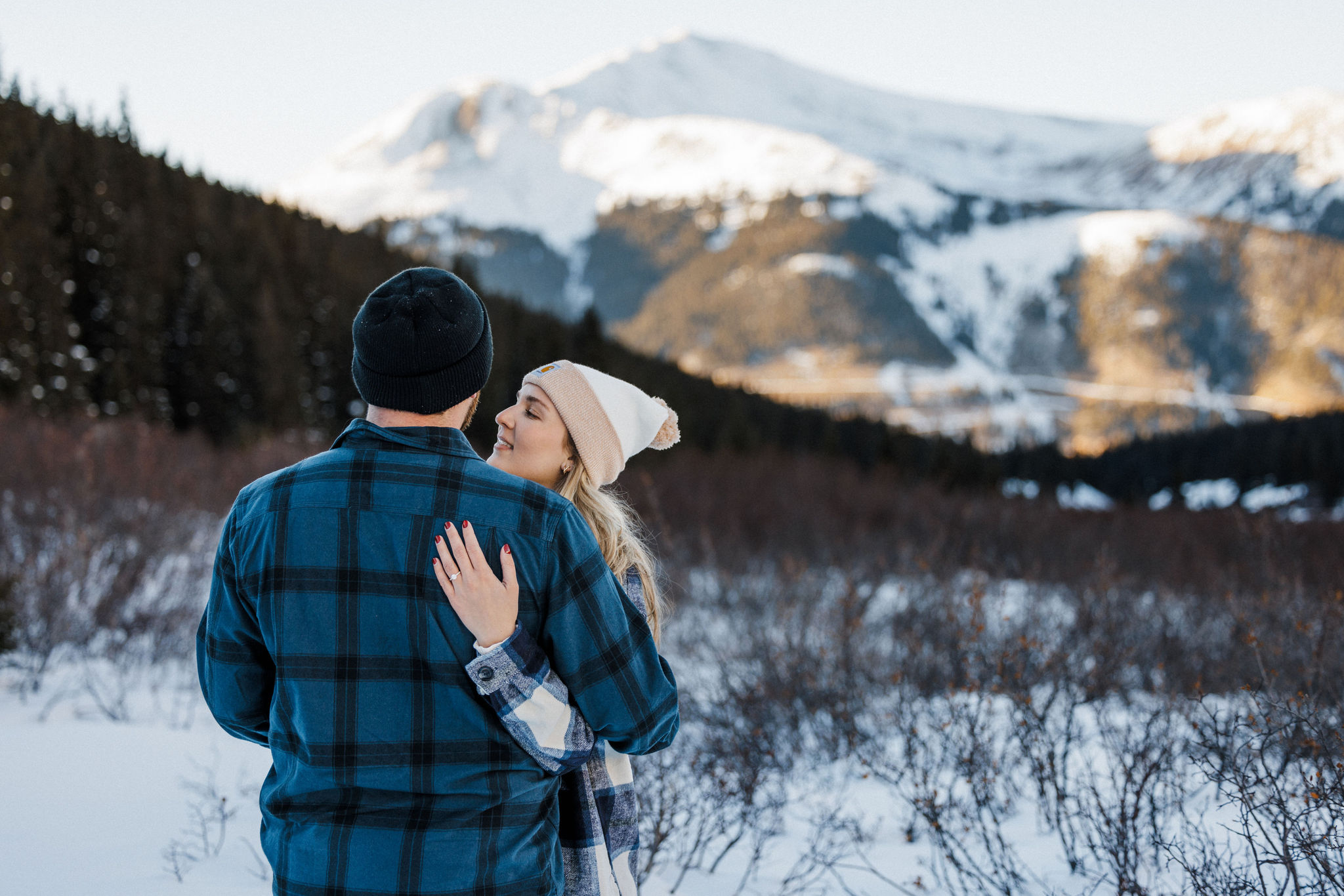 engaged couple smile at each other in front of the snowy colorado mountains