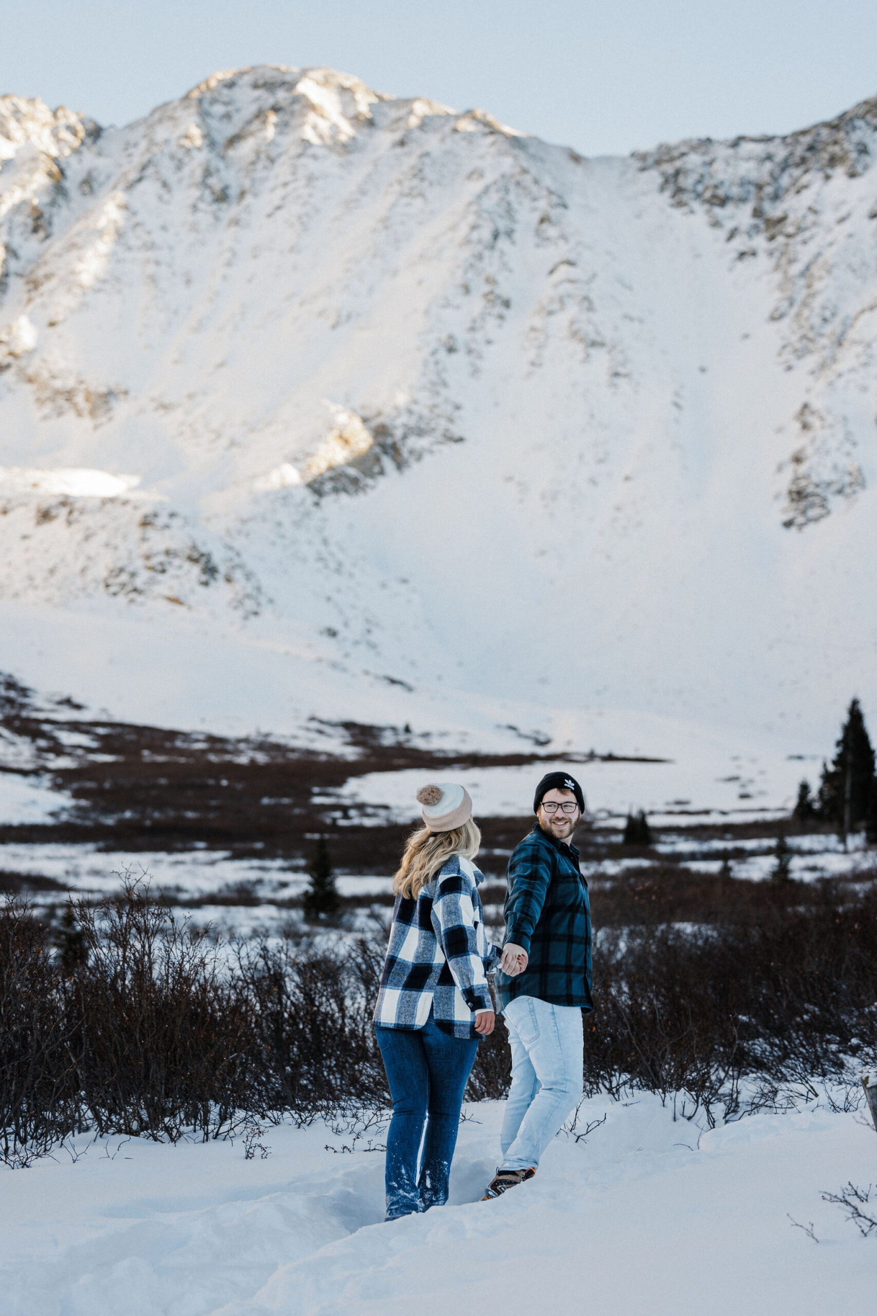 man and woman walk along snowy trail during winter engagement photos.