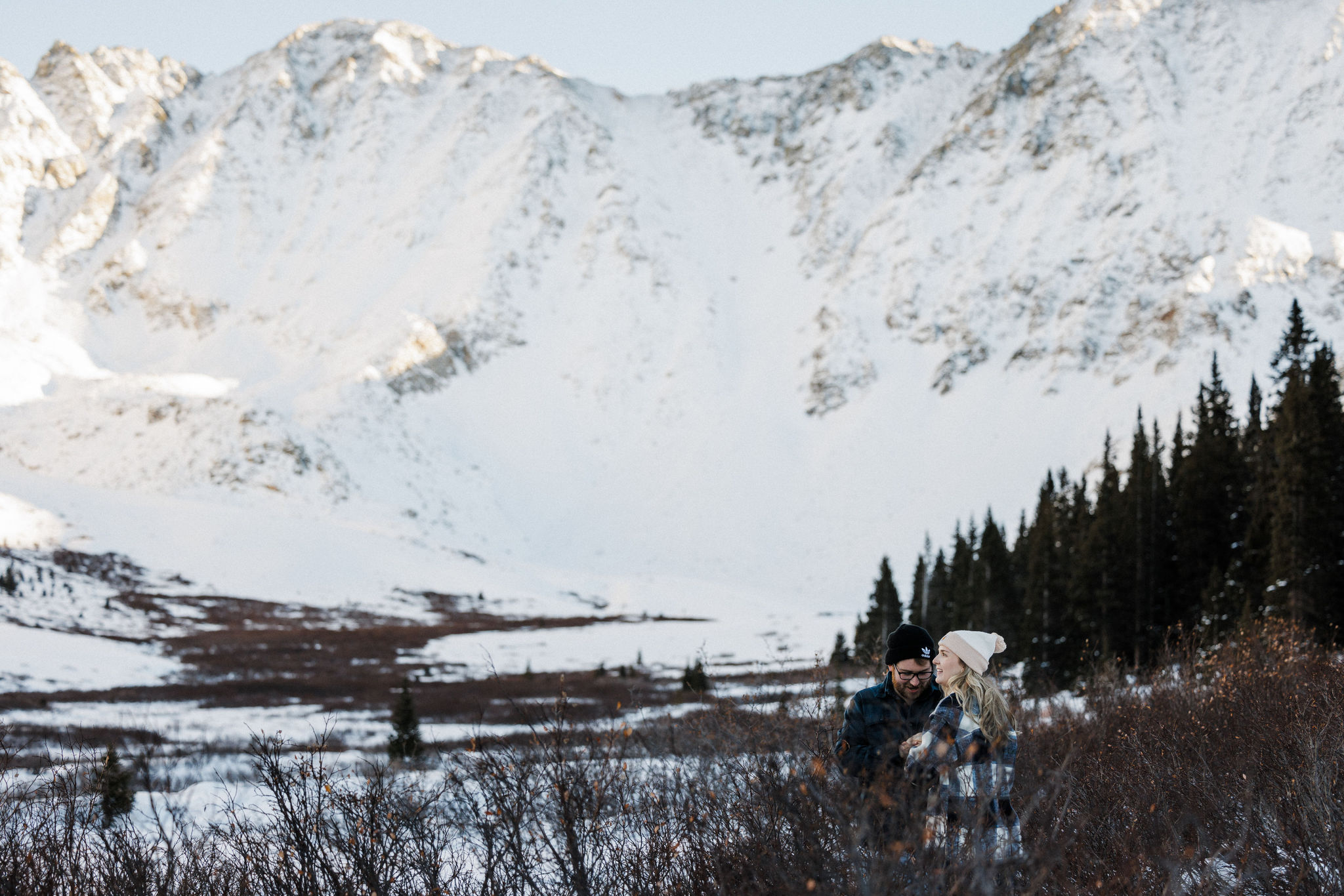 engaged couple stand in field under colorado mountains during engagement photos.
