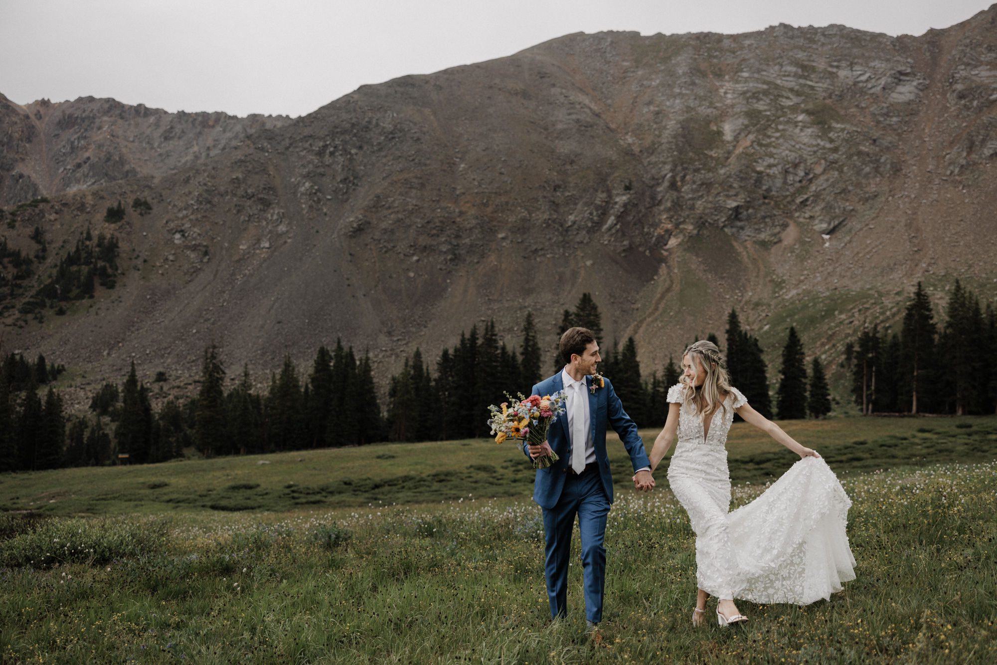 bride and groom hold hands as they happily walk through the fields below the mountains in colorado