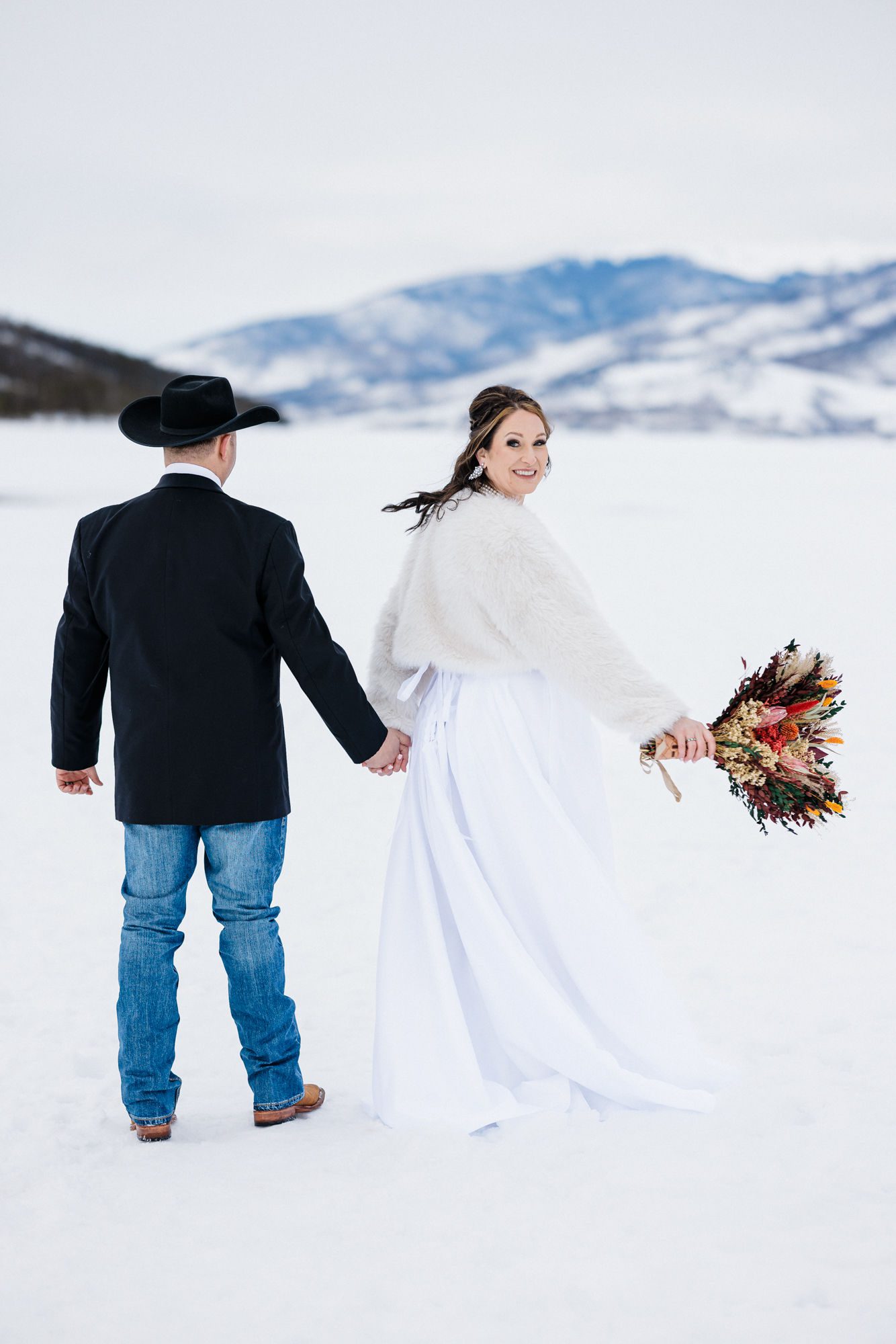 bride and groom hold hands and walk across frozen lake during winter elopement in colorado