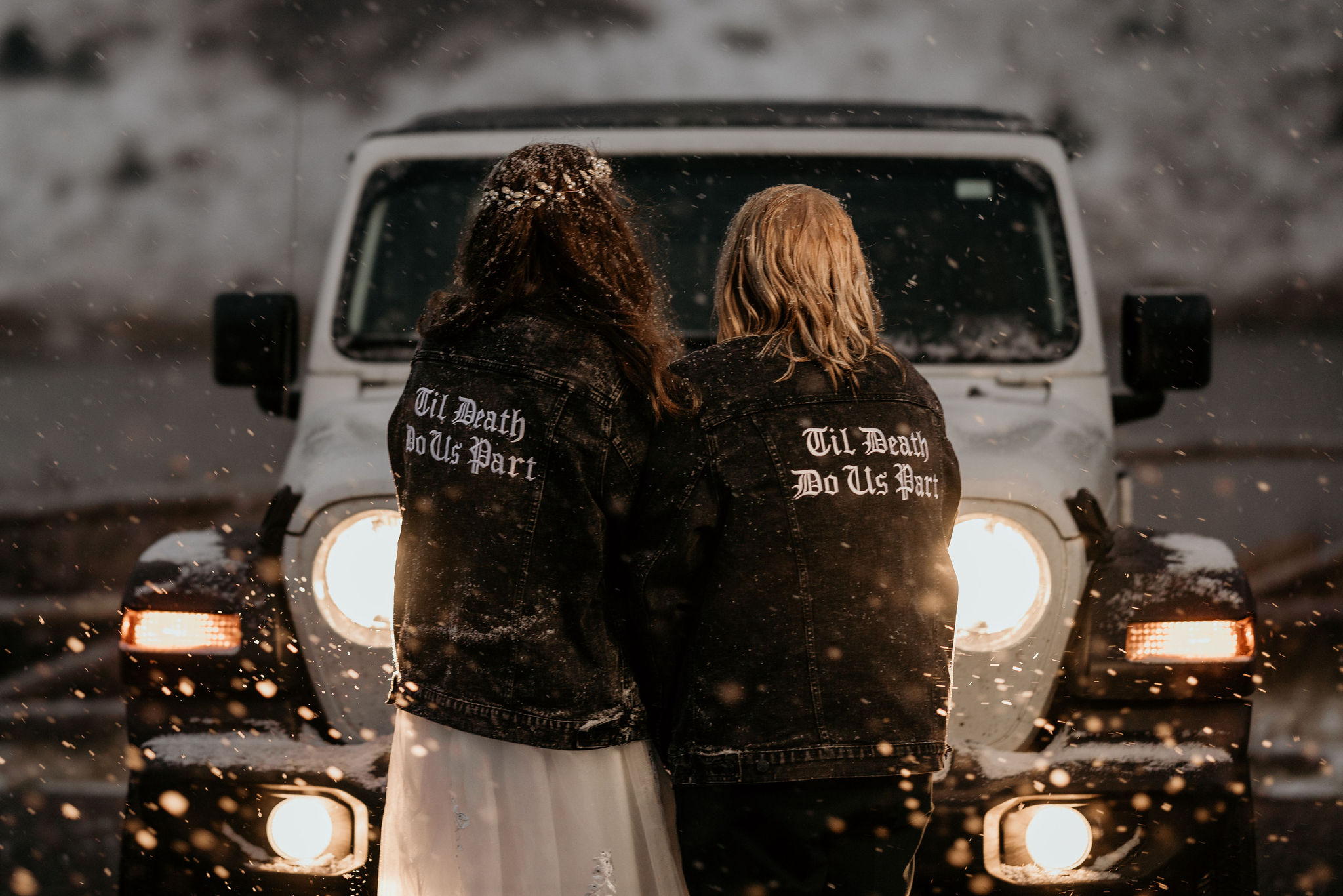two brides stand in the winter in front of a jeep with jackets that say 'till death do us part'