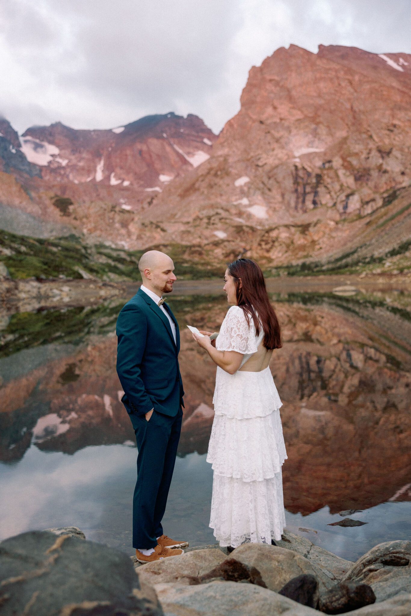 bride and groom stand on rock overlooking an alpine lake with mountains reflecting during their adventure colorado elopement package