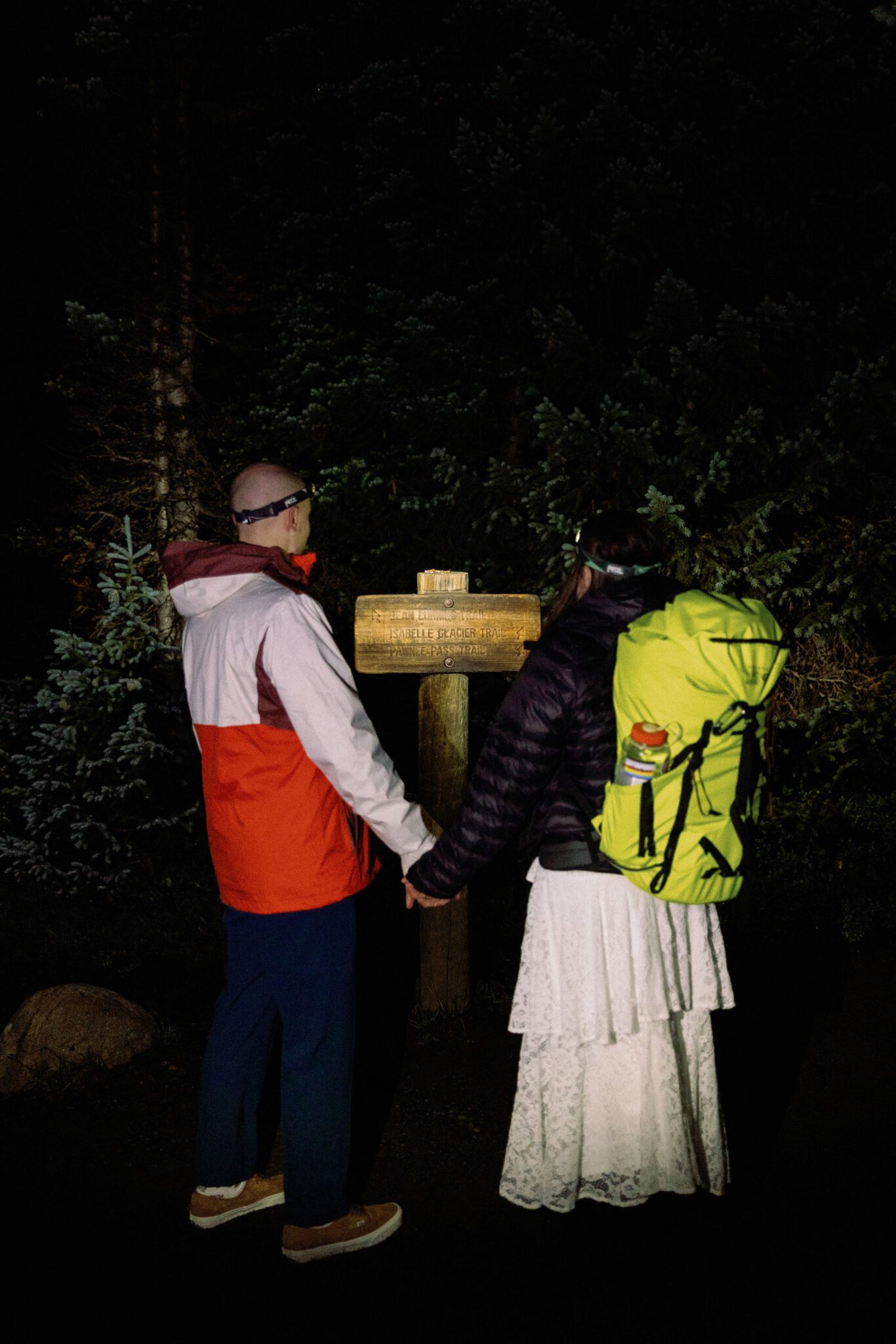bride with a hiking bag on her back and groom hold hands while looking at a trail sign during their elopement
