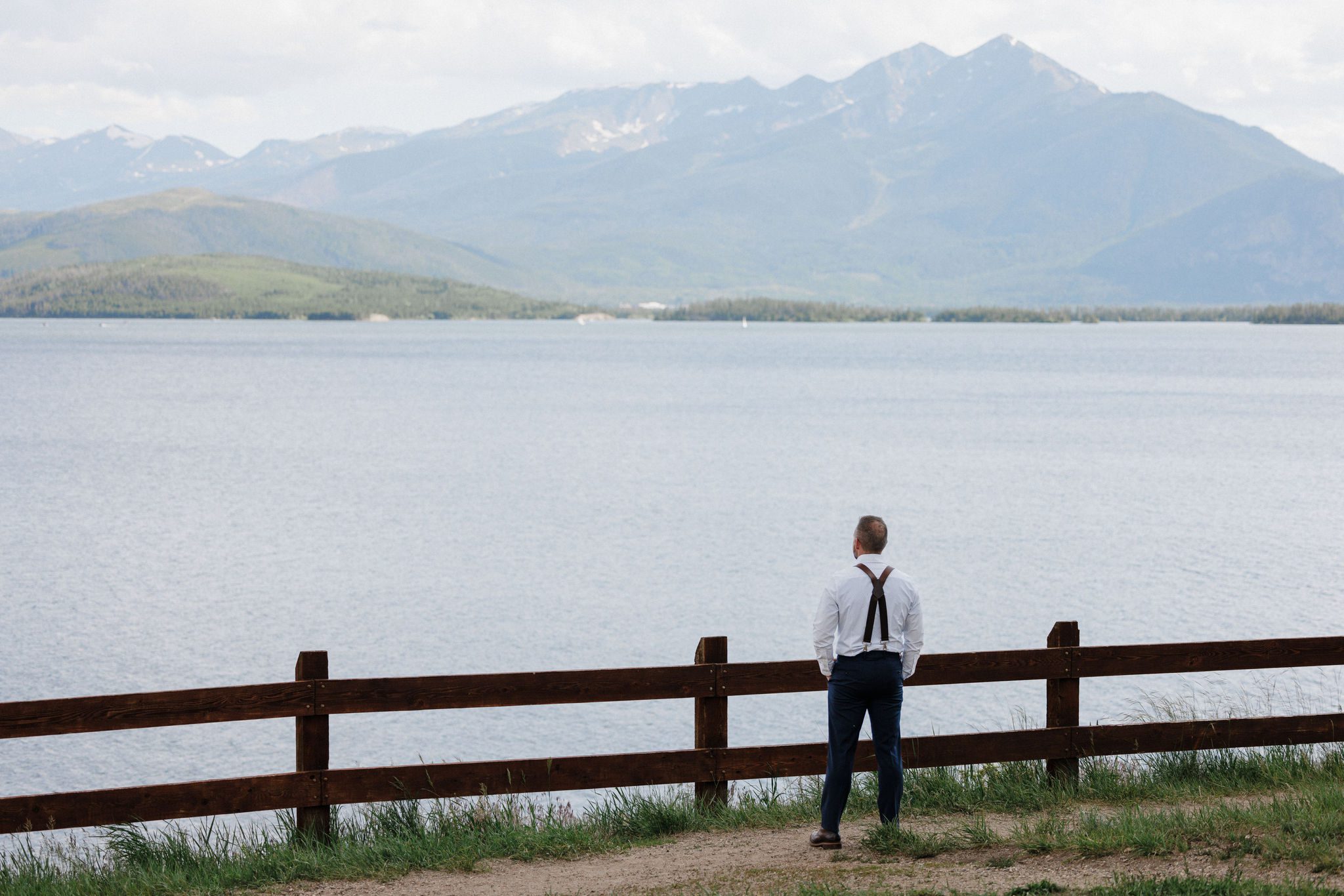 groom stands along a wooden fence and faces the mountain lake before their wedding first look