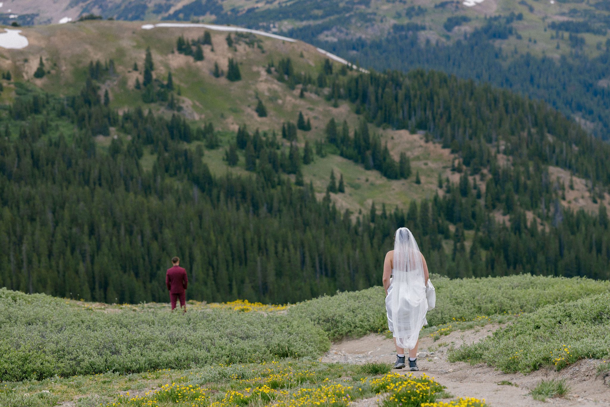 bride stands at the top of a hill in the mountains as the groom is at the bottom as a first look idea for their wedding