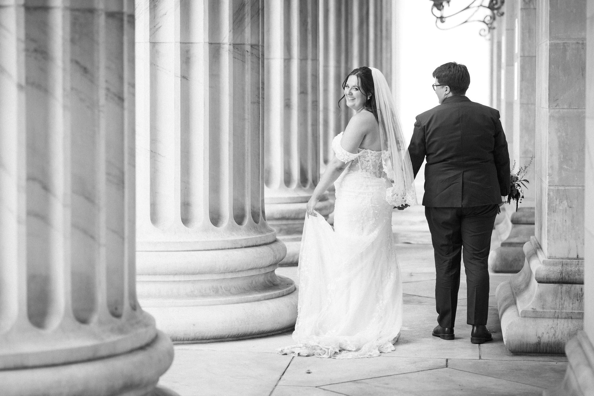 after telling family they eloped, two brides hold hands and walk through a marble courthouse
