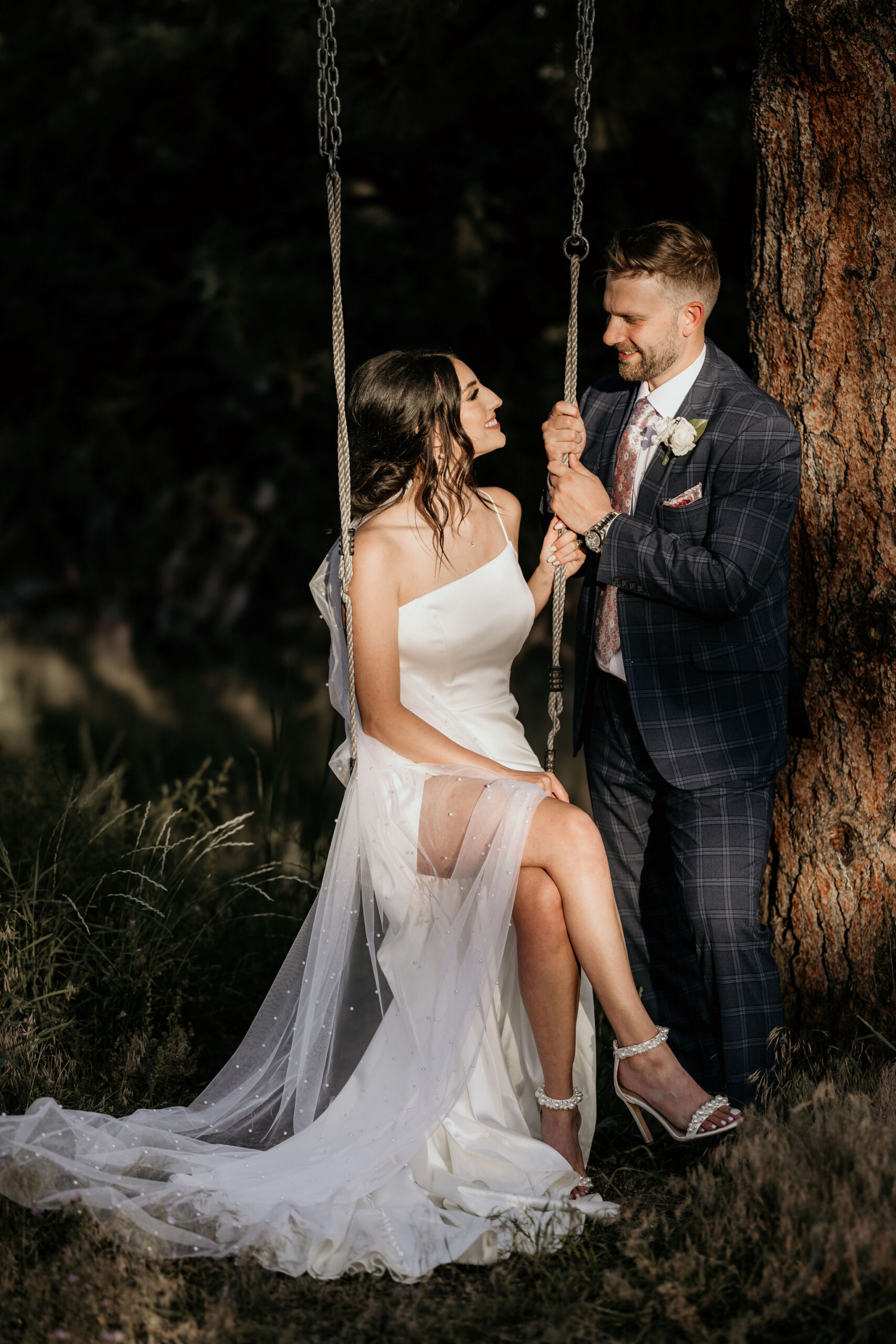 bride sits on tree swing and groom stands beside her during colorado elopement in an airbnb