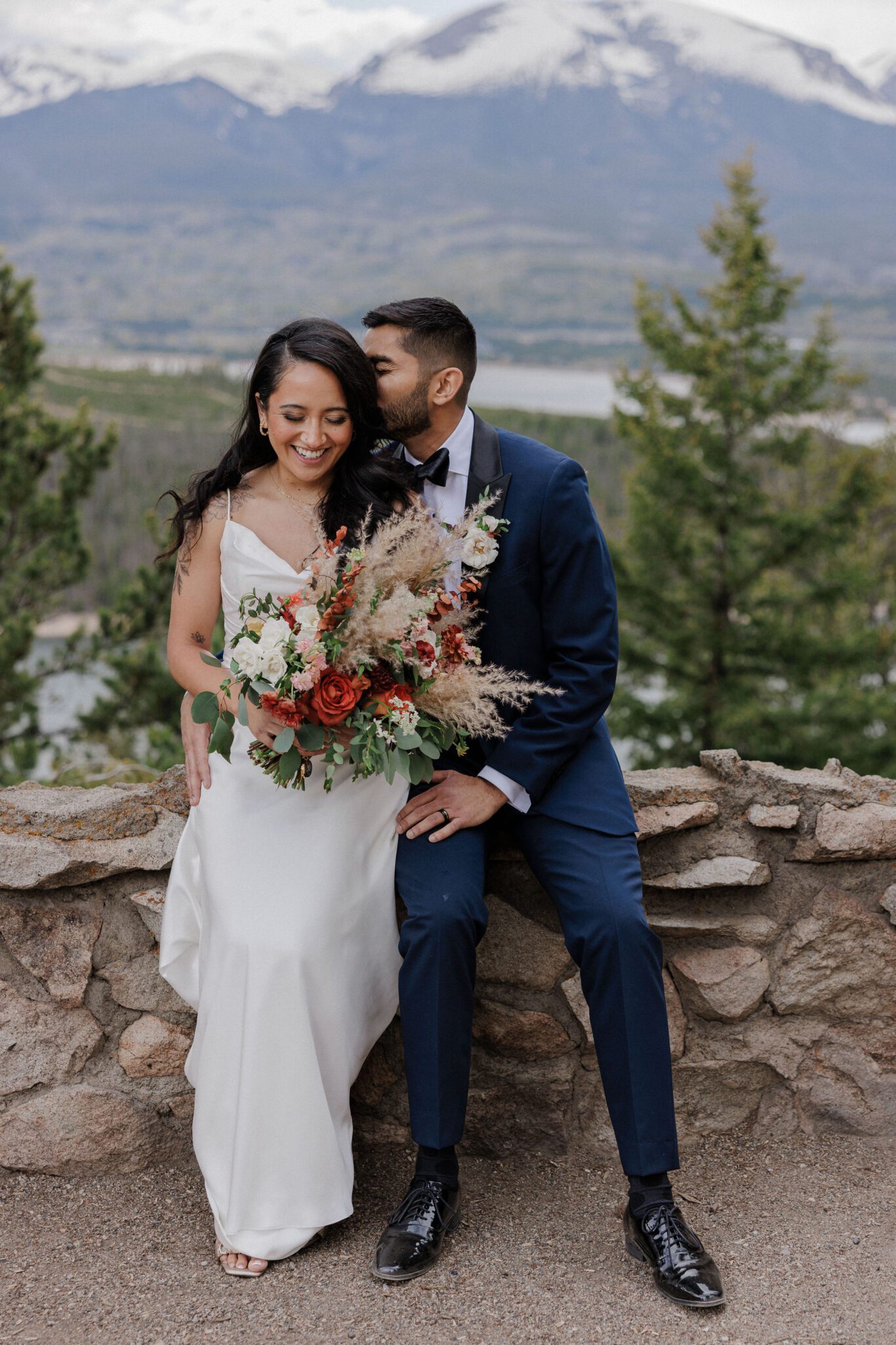 after telling family they eloped, bride and groom sit on rock ledge overlooking the mountains and a lake