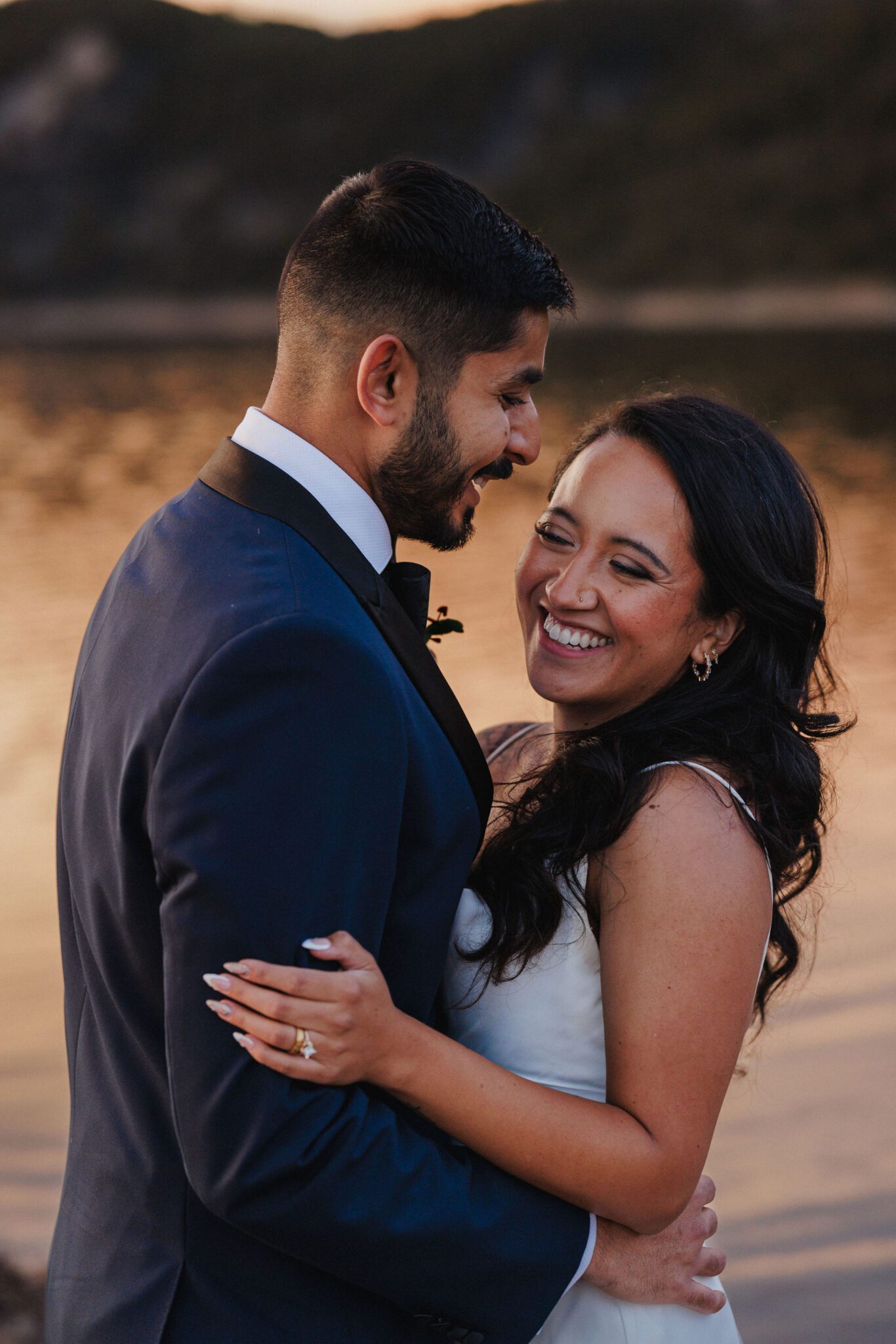 bride and groom embrace and smile at each other during sunset wedding photos by the lake