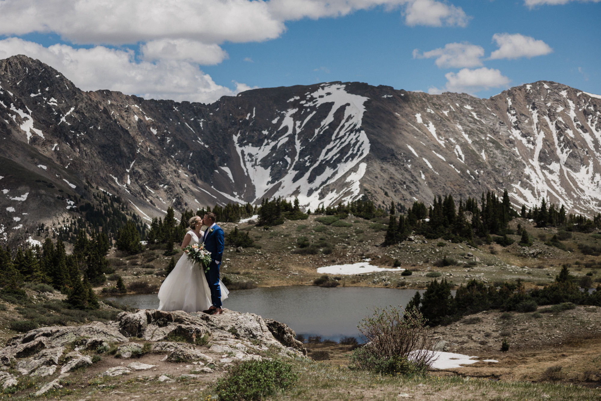 bride and groom stand on rock overlooking colorado mountains and lake on wedding day