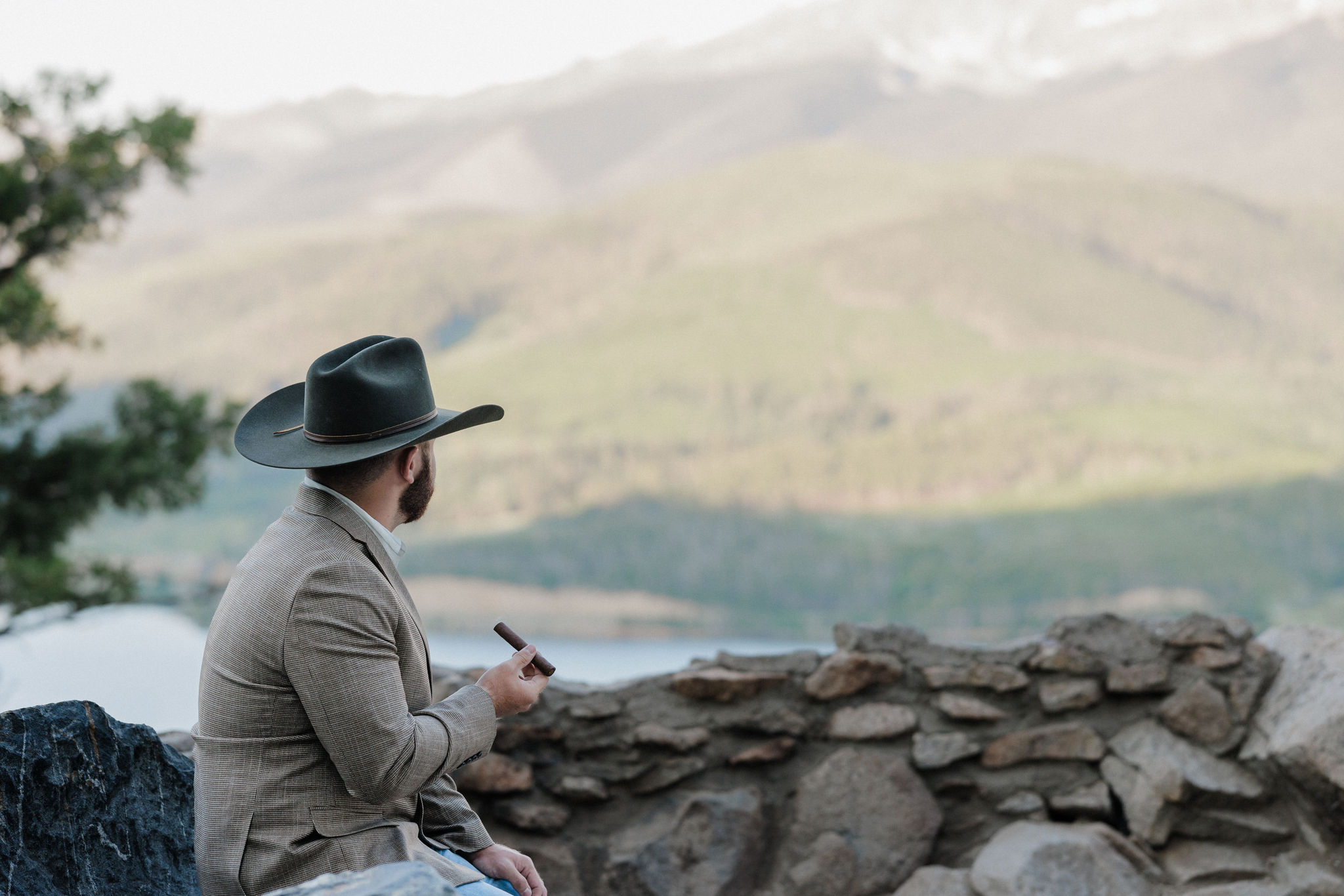 groom sits on rock and smokes cigar as he looks out at the mountains