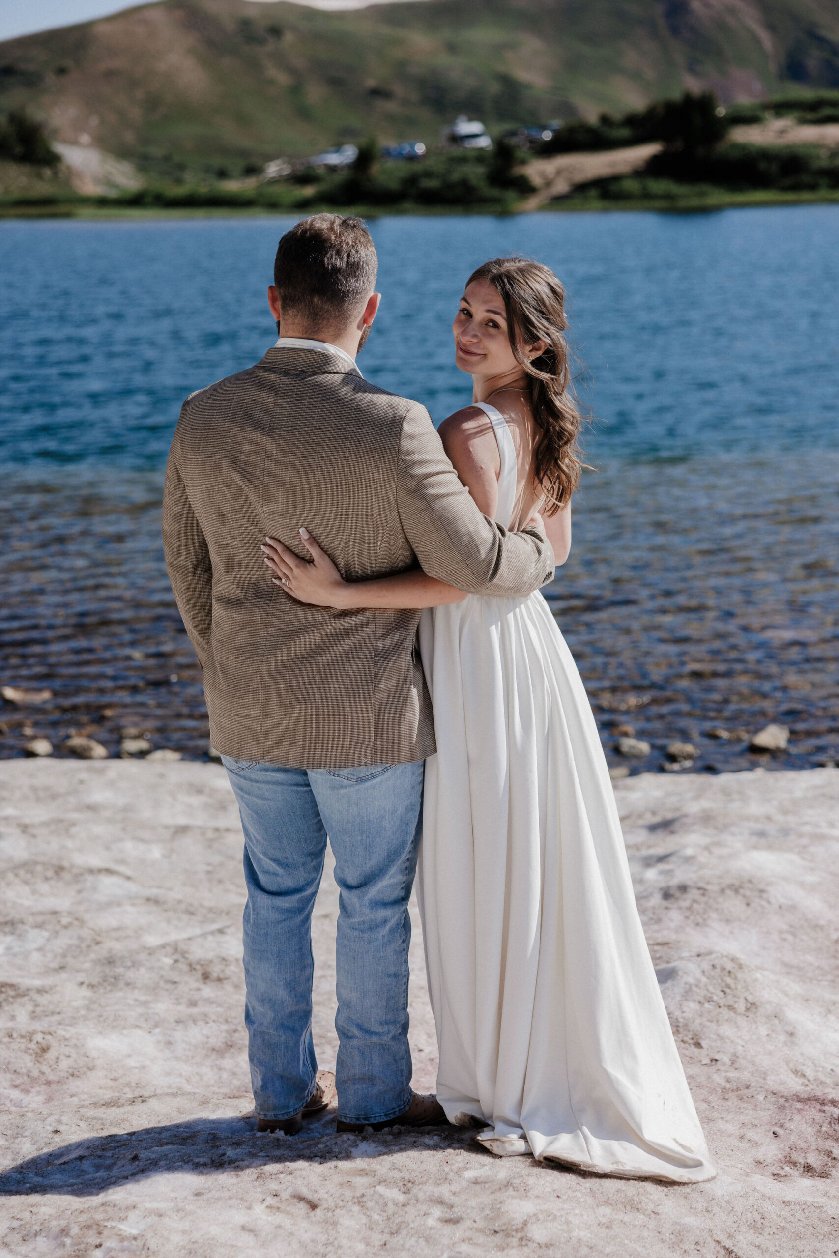 bride and groom stand in front of lake as bride looks back at elopement photographer