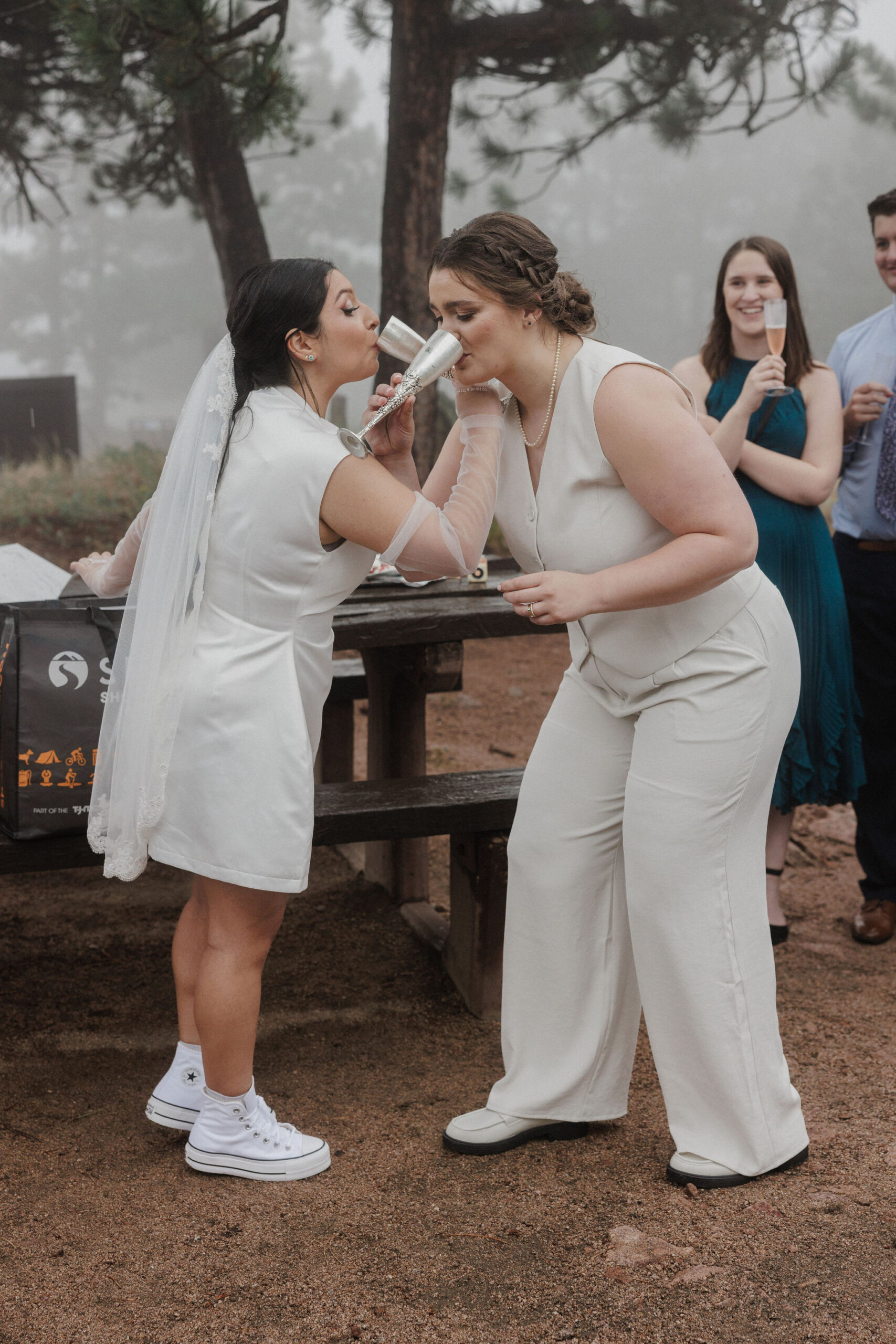 two brides interlace arms and sip glasses of champagne during mountain elopement