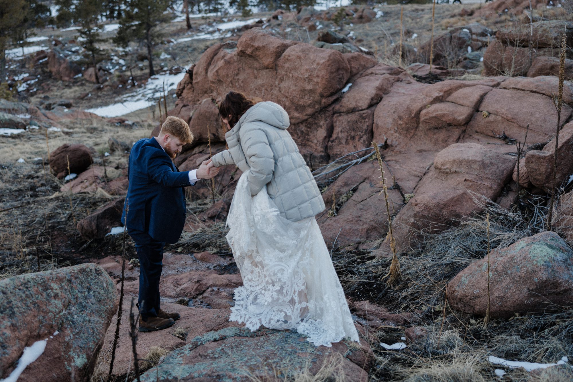 Groom helps bride walk down red rock formation at Airbnb elopement.