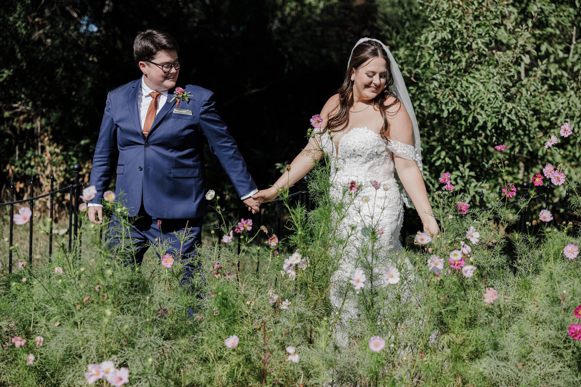 two brides hold hands and walk through flower field