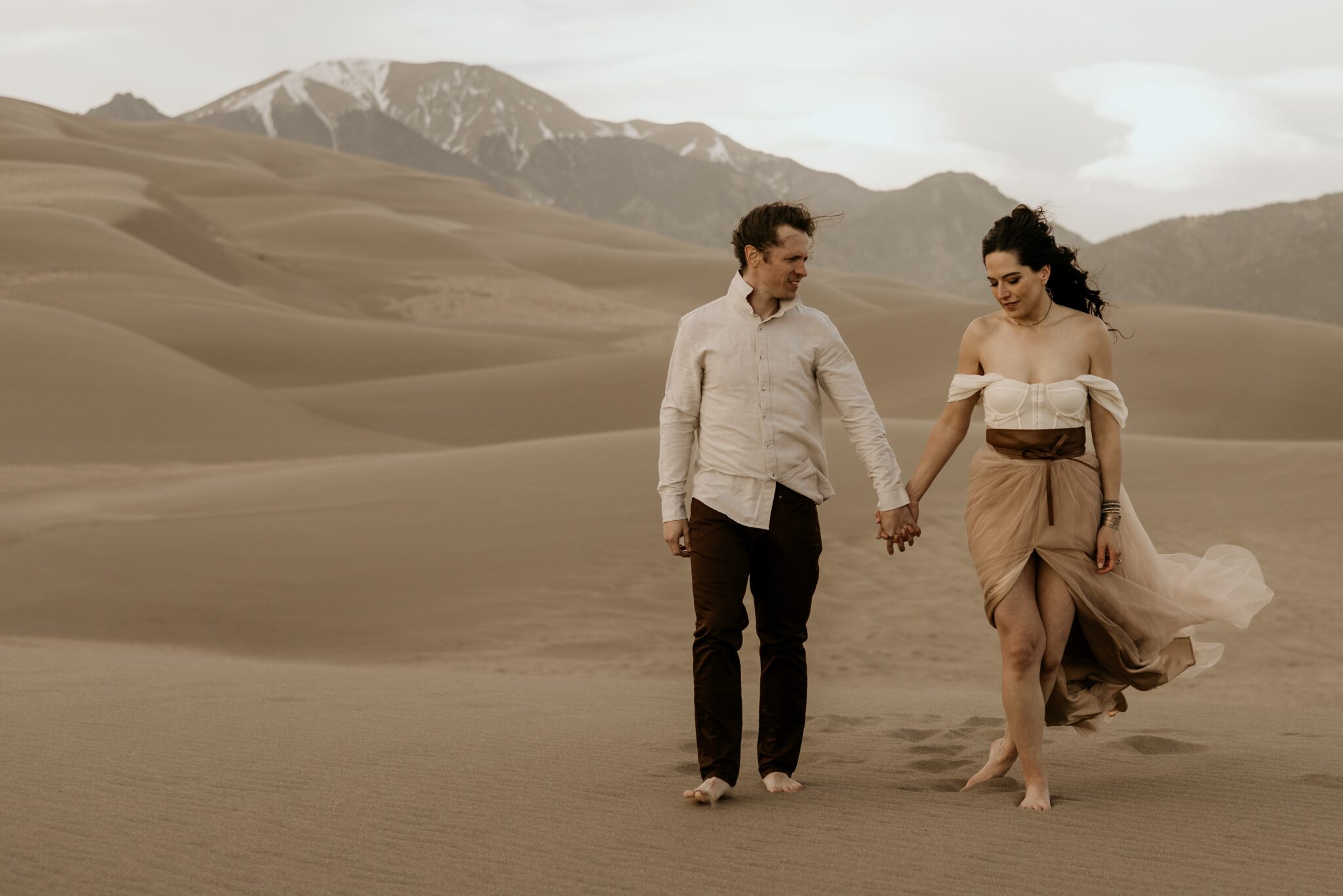 bride and groom hold hands while walking on the sand dunes, mountains in the background.