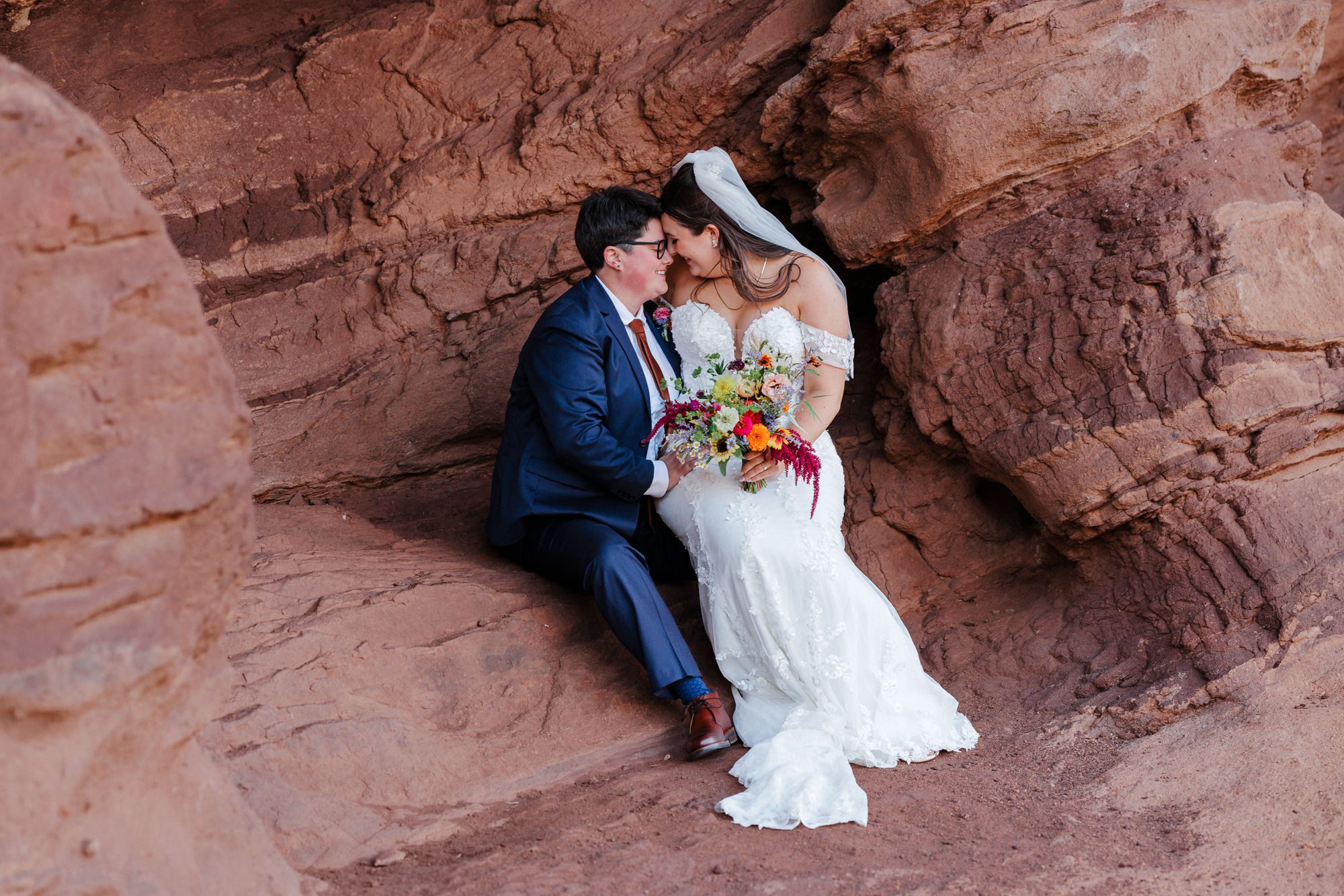 bride sits on the other bride's lap on a rock outcrop in Red Rocks Park during colorado elopement