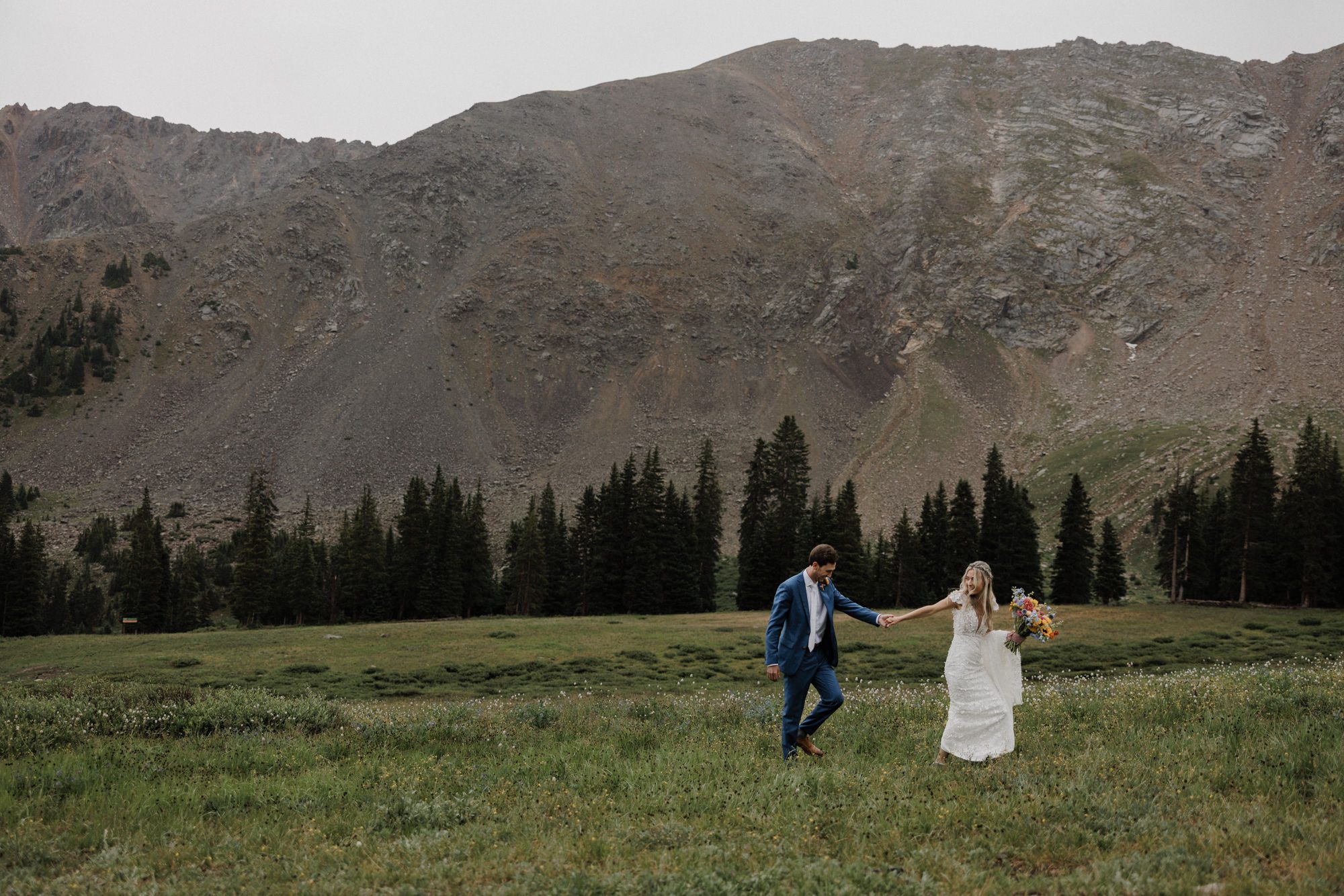bride and groom smile and hold hands as they walk through the field at the bottom of a mountain during their arapahoe basin wedding