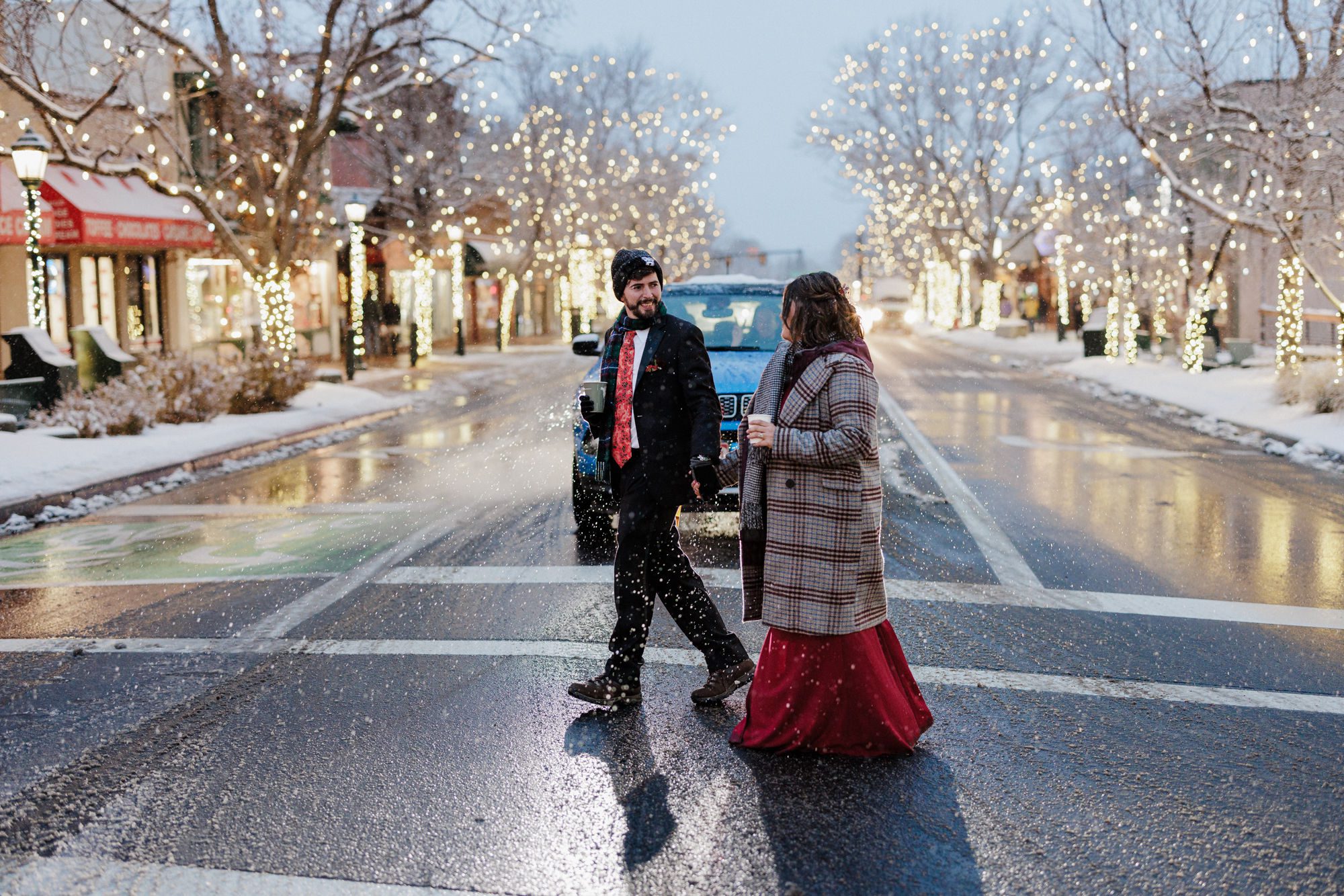 after choosing the winter colorado elopement package, bride and groom hold hands and walk across snowy street covered in holiday lights